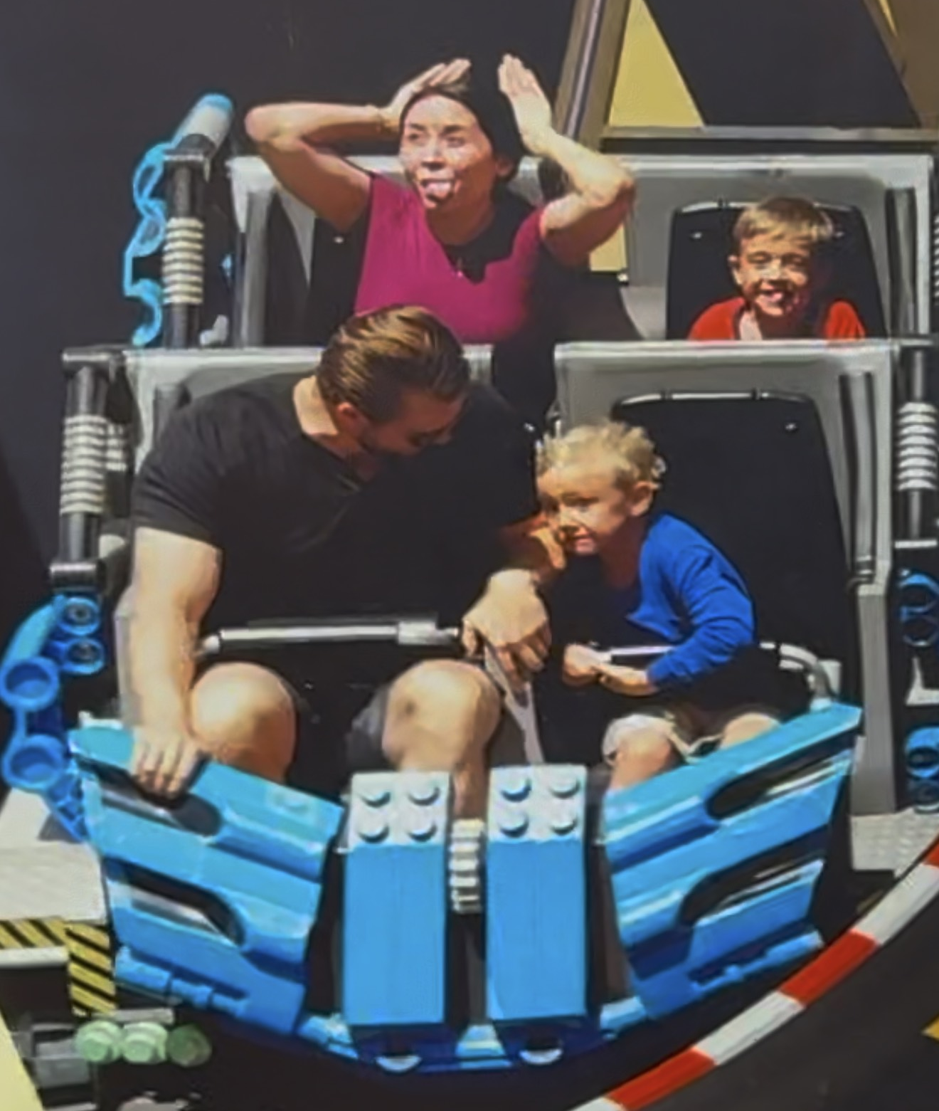 A family enjoying a ride on a LEGO-themed amusement park attraction. The boat-shaped ride is built with blue and black LEGO bricks, and the family members are sitting inside it, expressing joy and excitement.