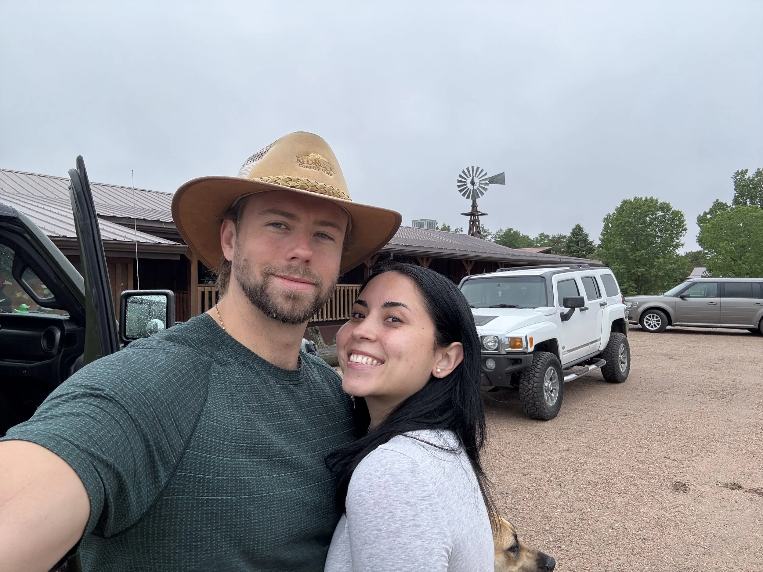 A young man wearing a cowboy hat and a young woman with black hair smiling for a selfie outdoors, with three parked vehicles and a rustic building in the background, along with a windmill and trees.
