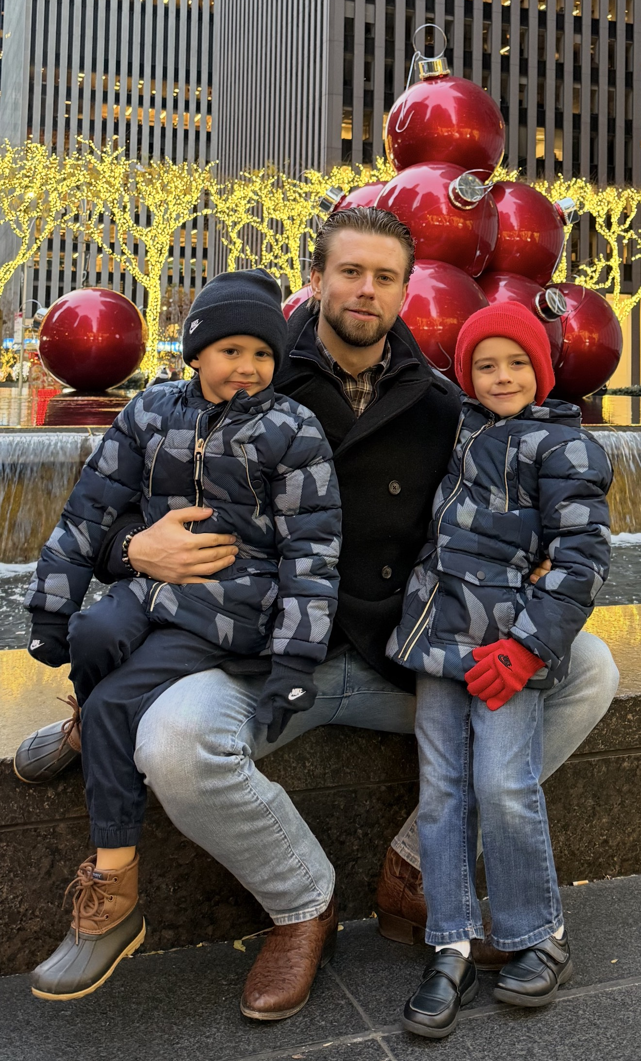 A man and two children sitting on a fountain in front of large red Christmas ornaments and yellow-lit trees in an urban area.
