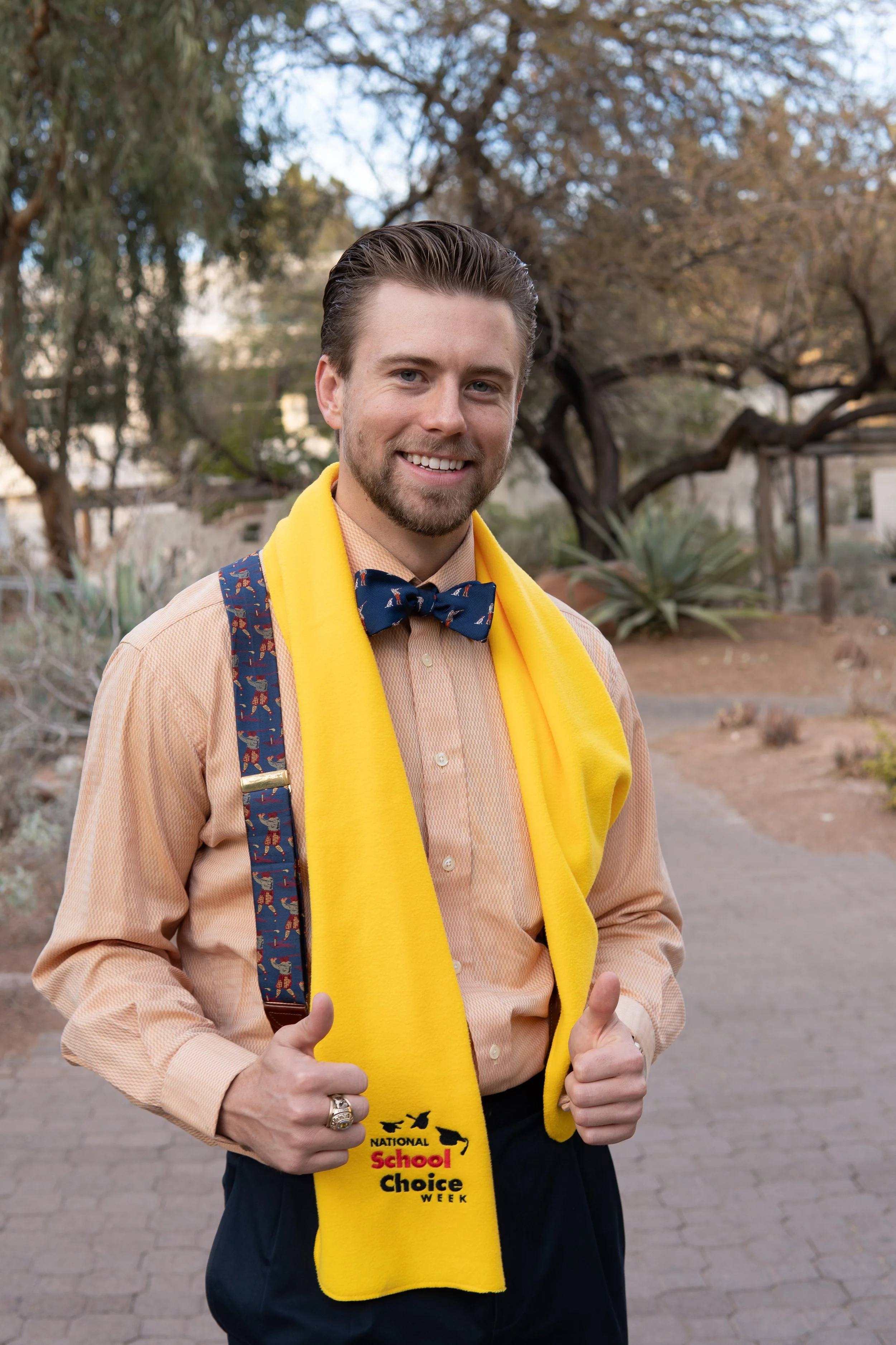 A man smiling outdoors wearing a yellow scarf with 'National School Choice Week' logo, a navy bow tie, peach shirt, and suspenders with a pattern, giving a thumbs-up.