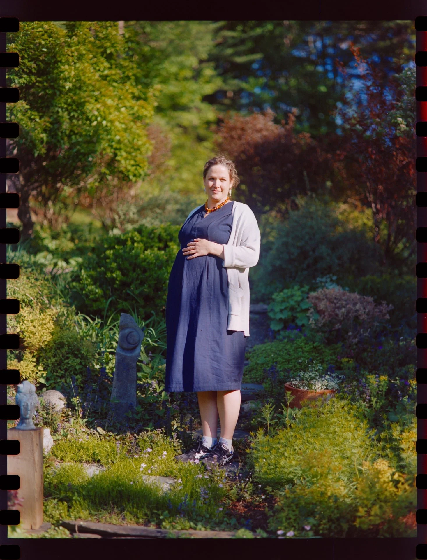 A woman standing in a lush garden with green plants and colorful trees in the background, wearing a dark dress, light sweater, and sneakers, smiling and holding her chest with one hand.