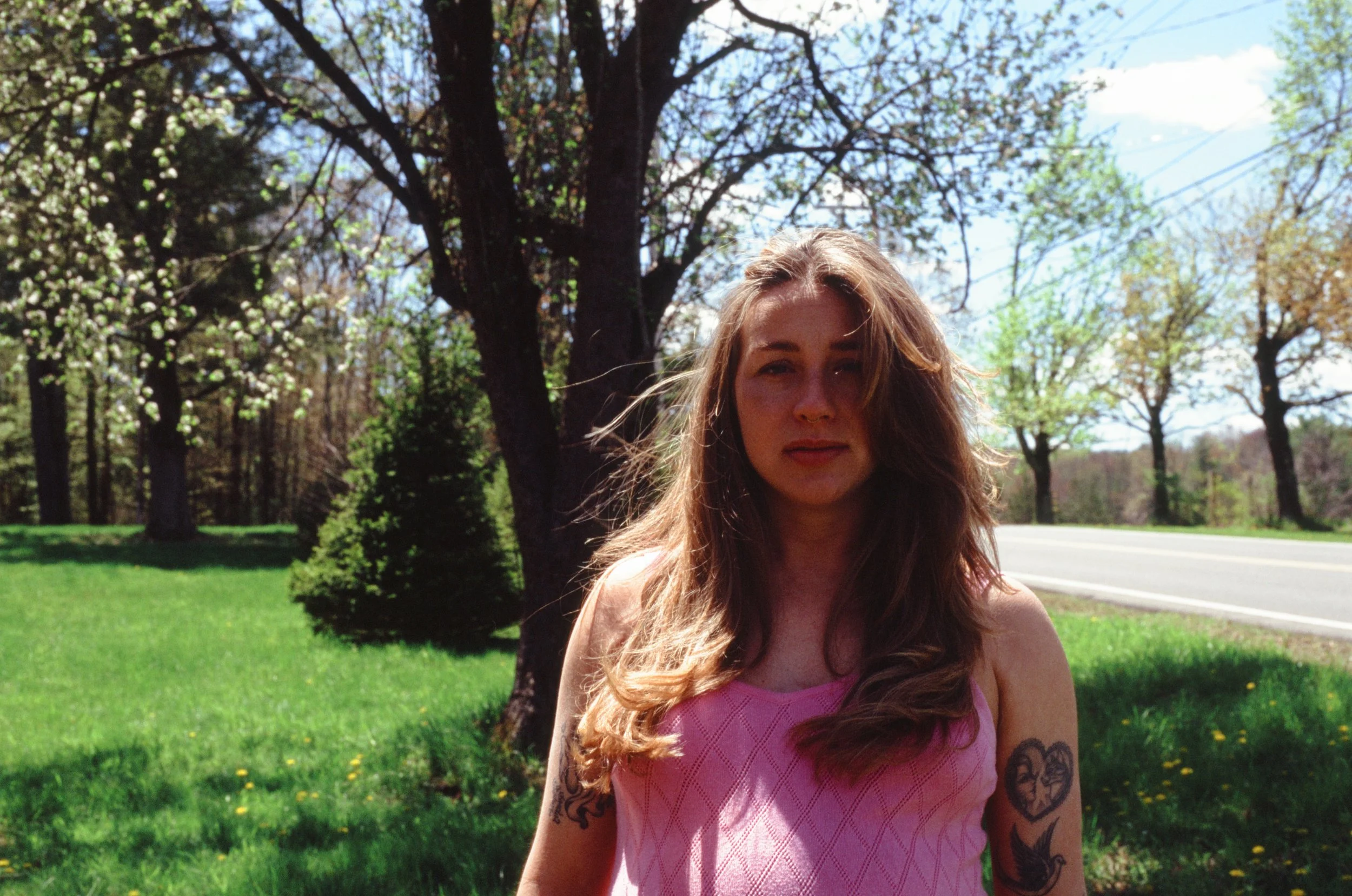 A woman with long red hair and tattoos on her arm wearing a pink top outdoors among trees and greenery on a sunny day.