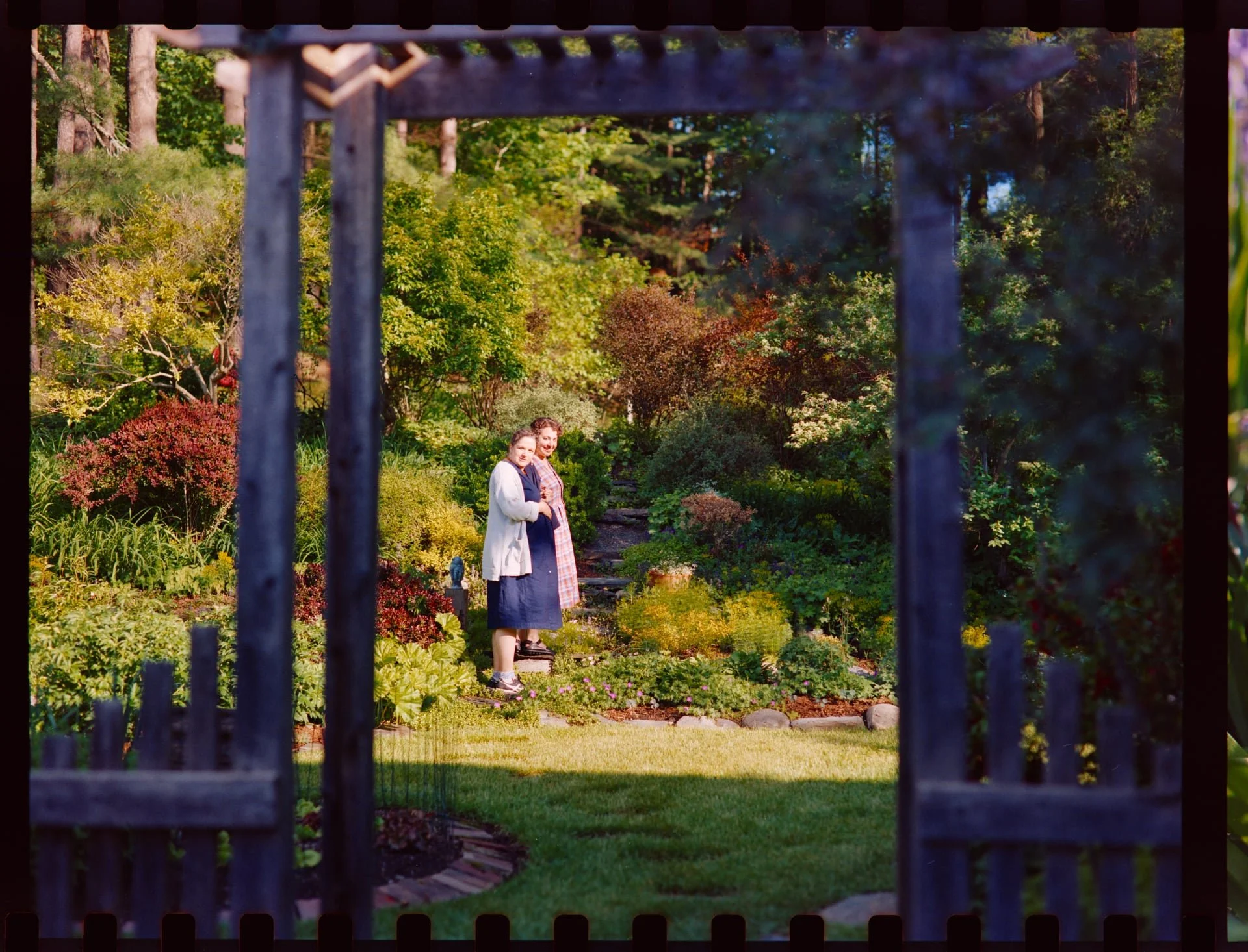 Two women standing in a lush garden with various shrubs and trees, seen through black wrought-iron fence.