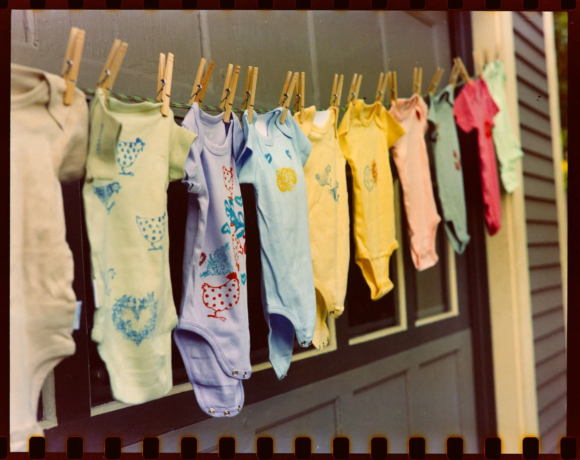 Multiple pastel-colored baby onesies hanging on a clothesline with clothespins in front of a brown wooden wall.