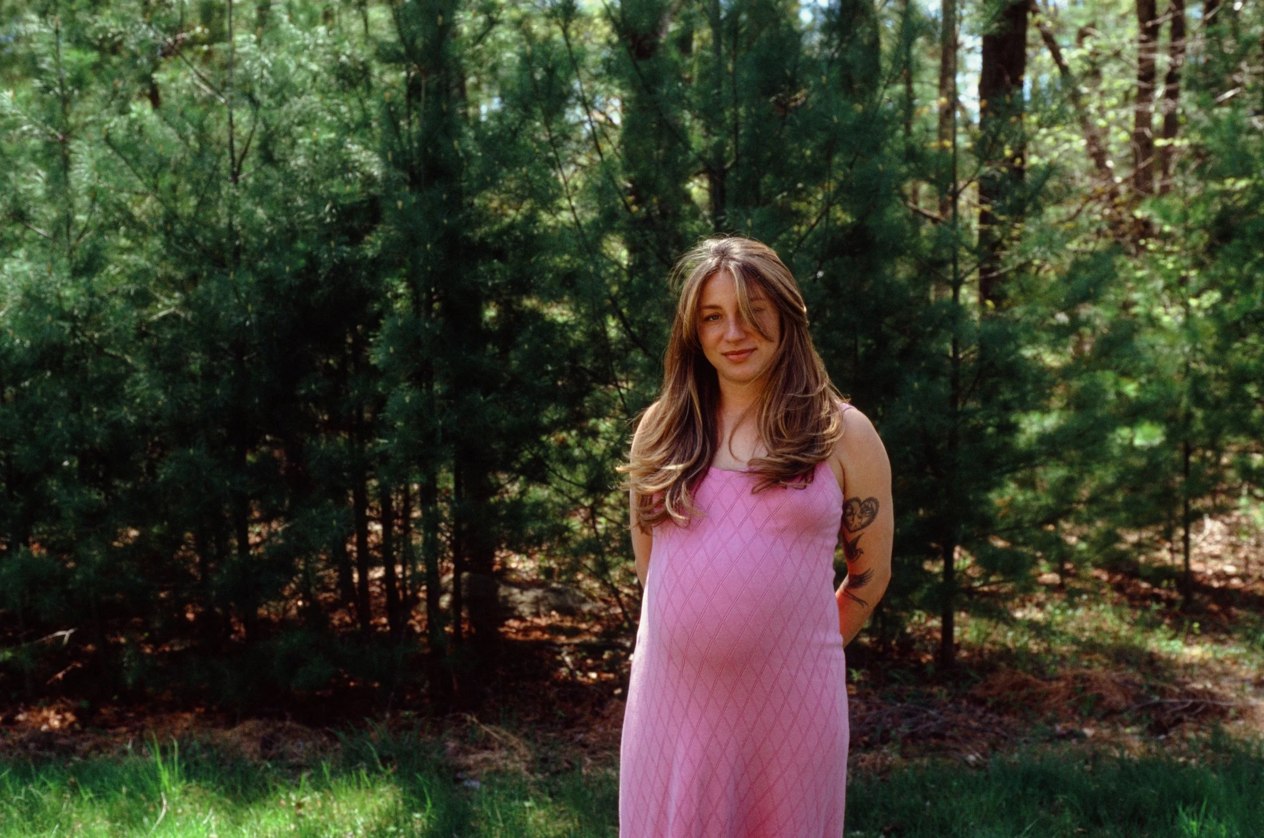 A woman with long wavy blonde hair wearing a pink dress with diamond patterns stands outdoors in front of a background of green trees and foliage, smiling softly. Maternity photograph. 