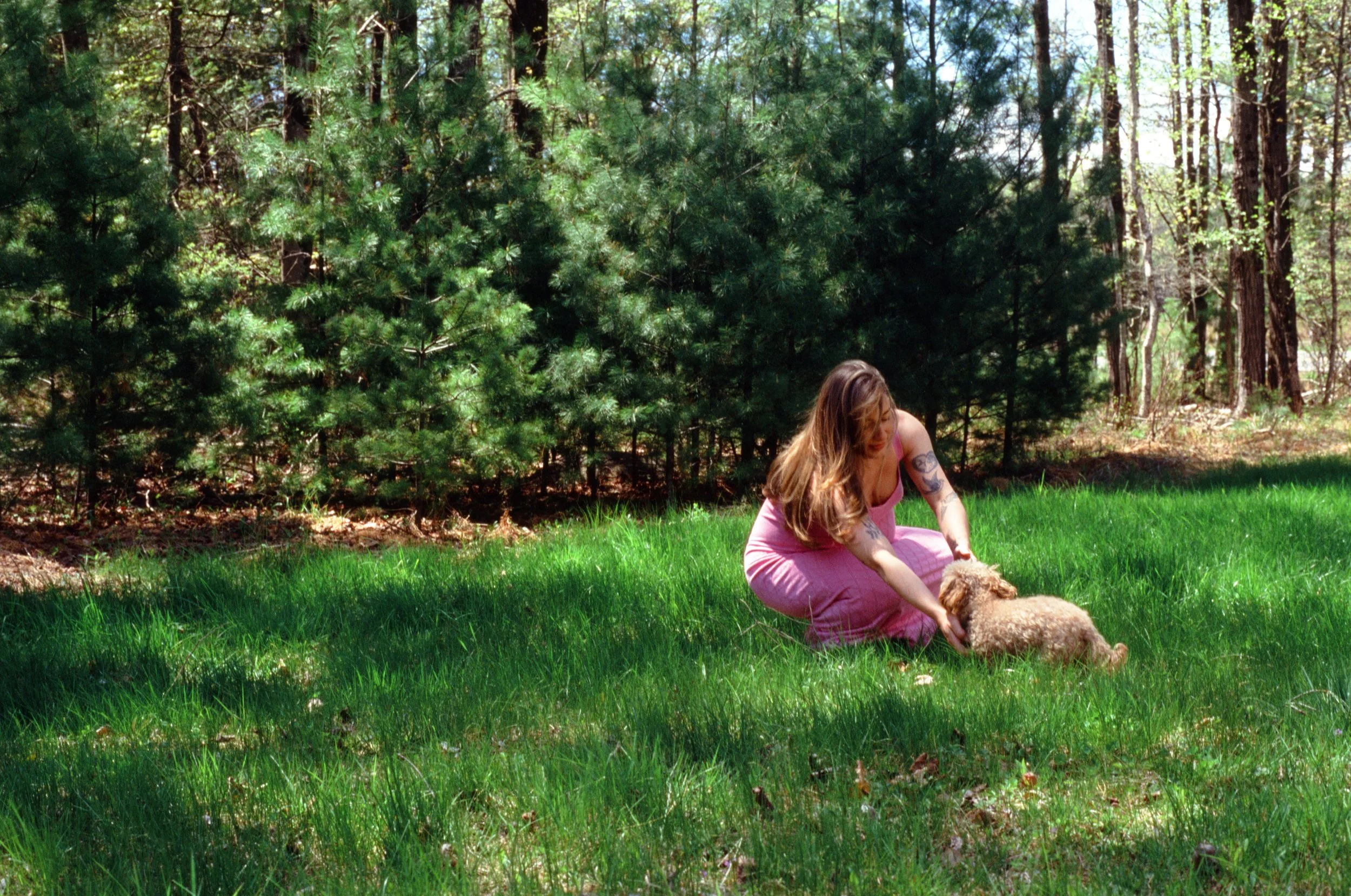 A woman with long hair sitting on the grass in a forested area, playing with a light-colored dog.