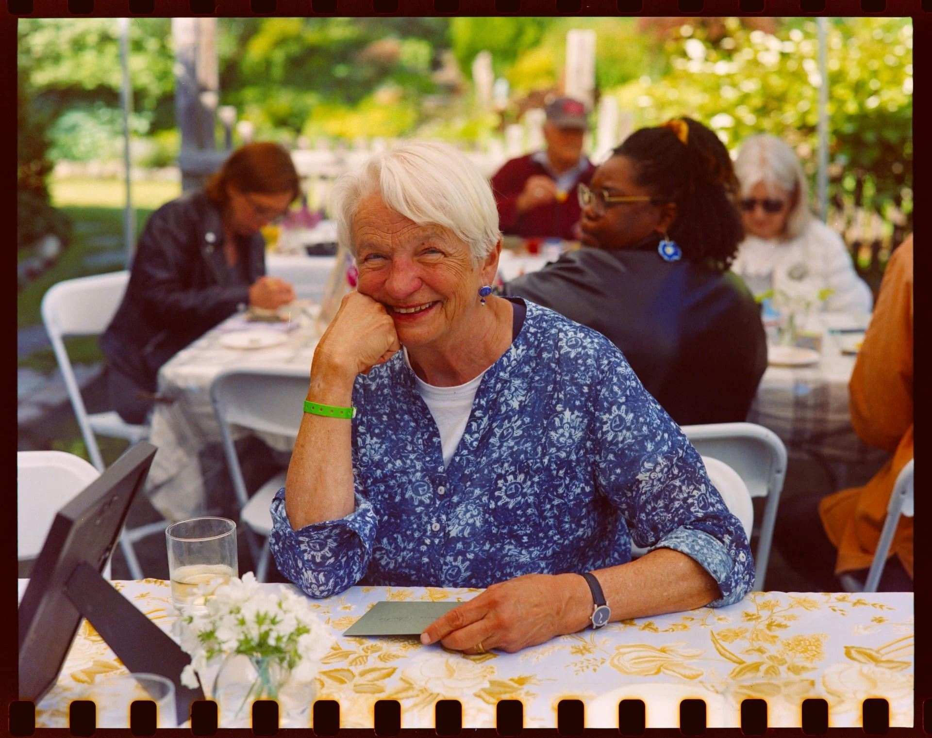 An elderly woman with white hair and a blue patterned shirt is smiling at a gathering outdoors. She is sitting at a table with a yellow tablecloth, flowers, and drinks. Other people are seated around her, and the setting appears to be a garden or par