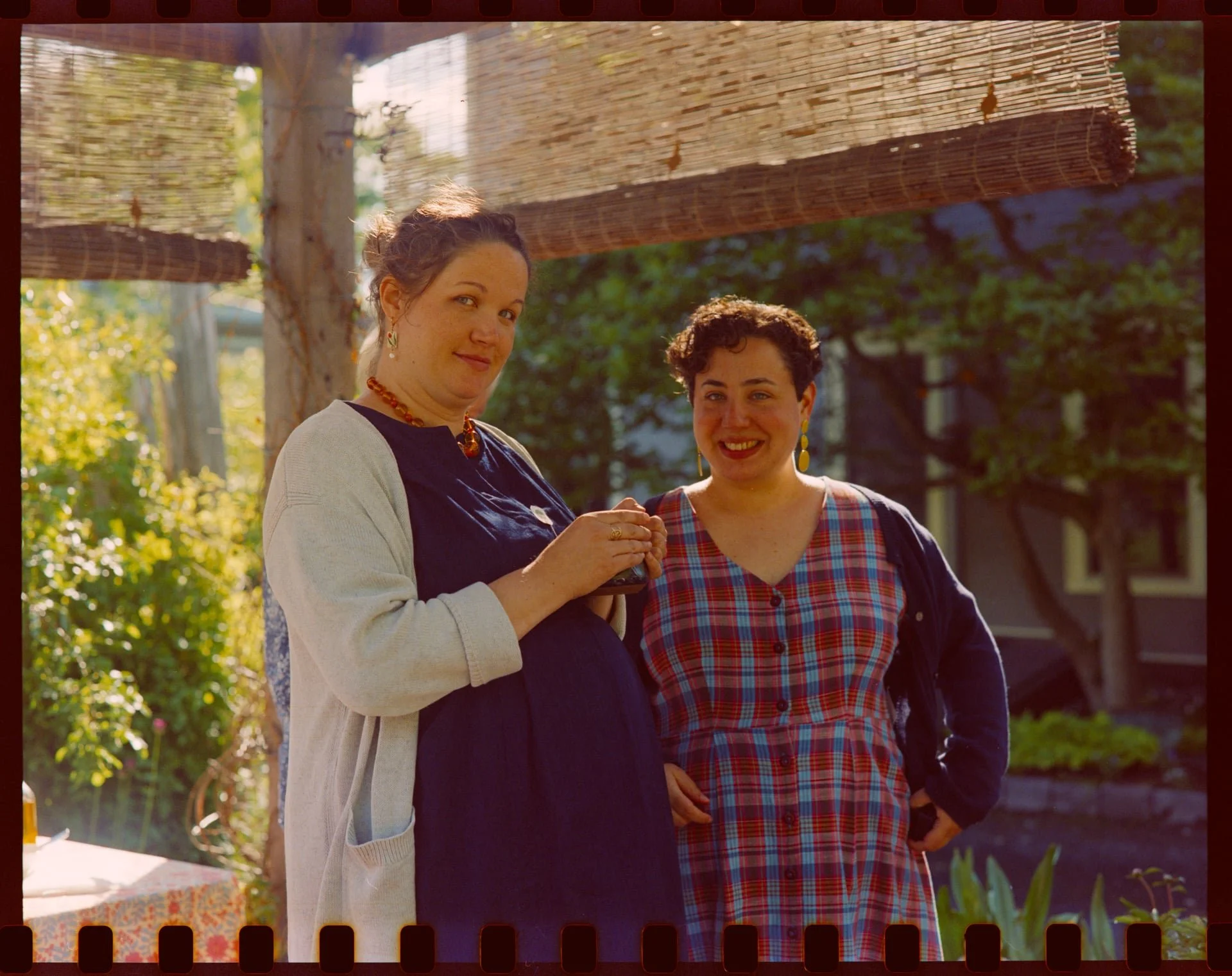 Two women standing outdoors in a garden or park area, smiling and holding hands, with a hanging bamboo shade overhead.