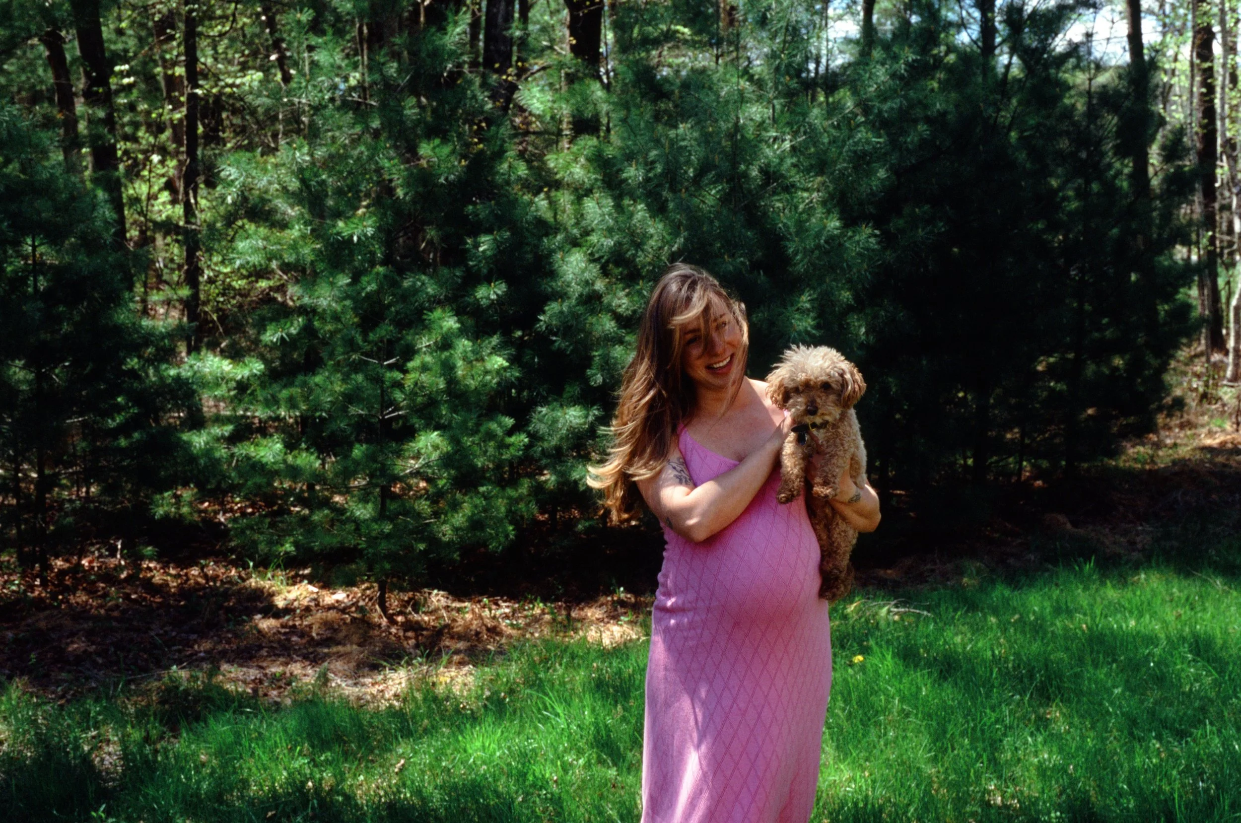 A smiling woman with long hair holding a small brown poodle dog in a forested area with trees and grass.
