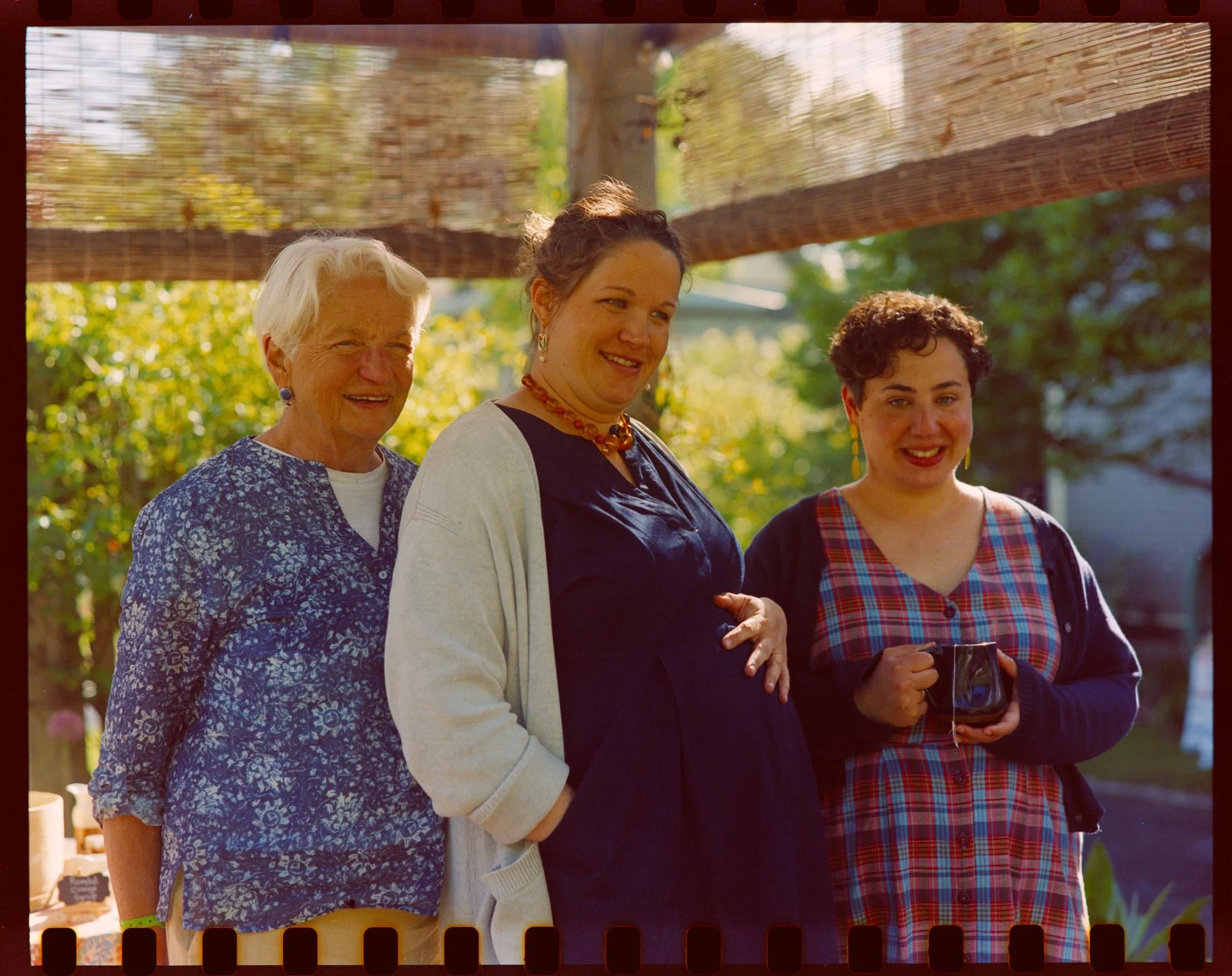 Three women smiling outdoors, standing close together with one holding a glass of wine, under a bamboo shade, with trees in the background.
