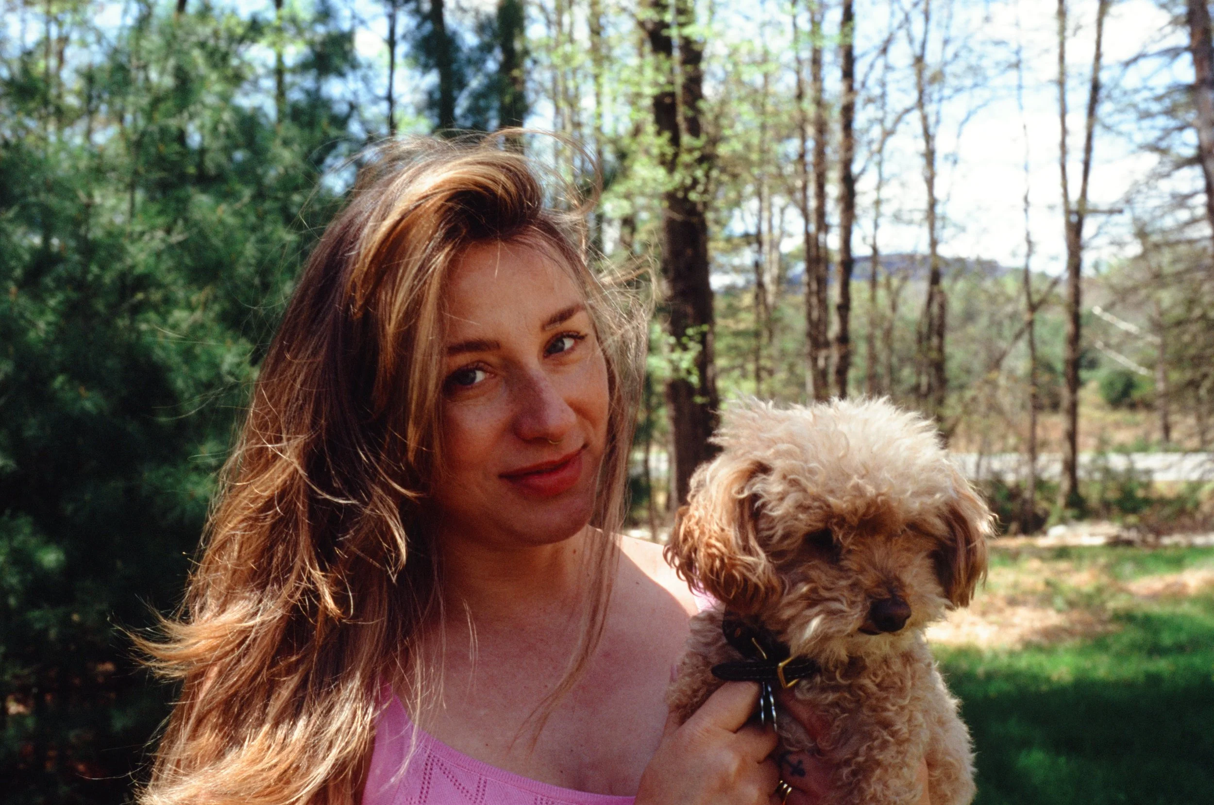 A woman with long, wavy about reddish-bopper hair holding a small, curly-haired dog outdoors in a wooded area with tall trees and sunlight.