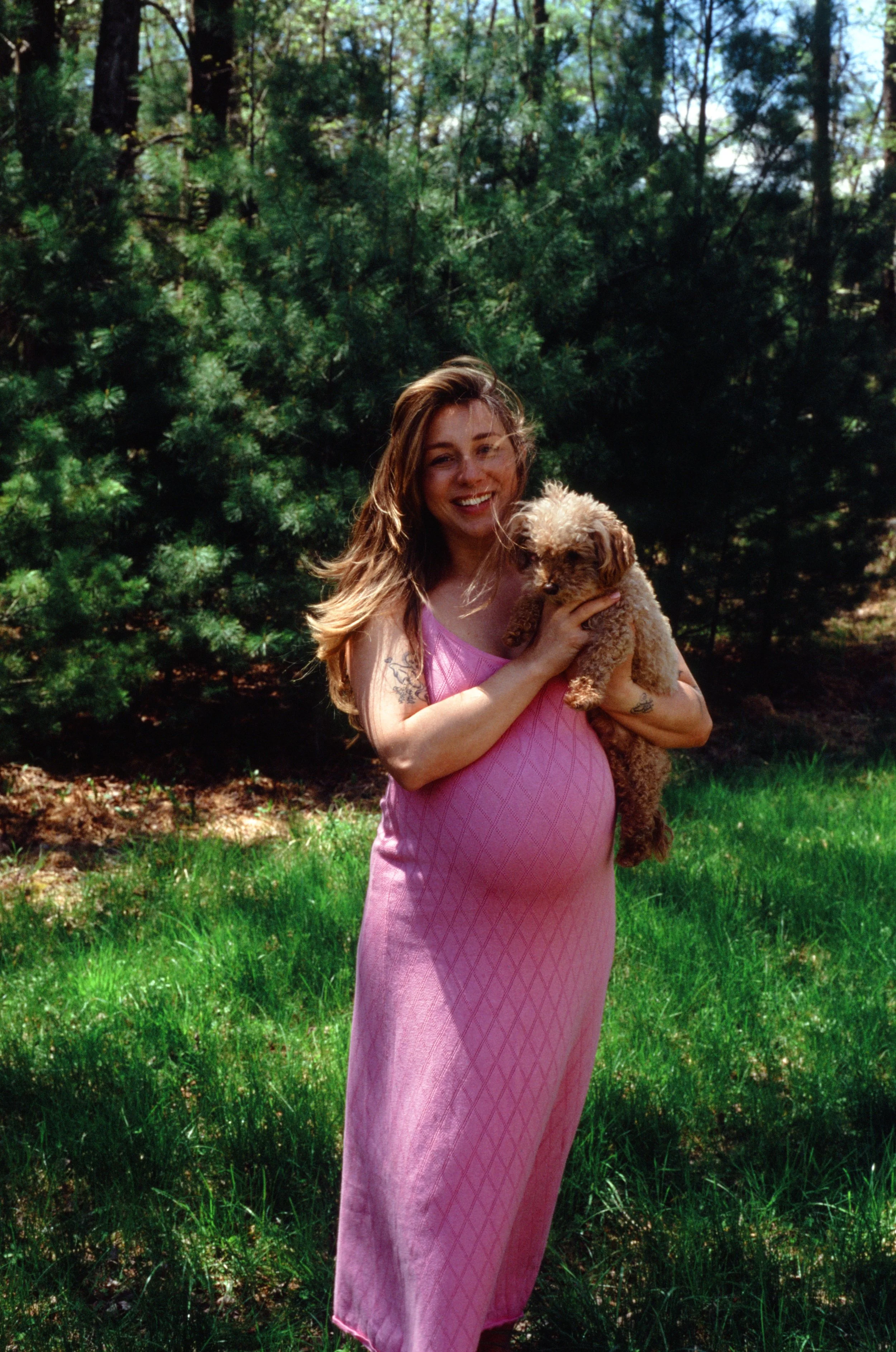 A woman with long, wavy hair holding a small, curly-haired dog outdoors in a grassy area with trees in the background. She is smiling and wearing a pink dress.