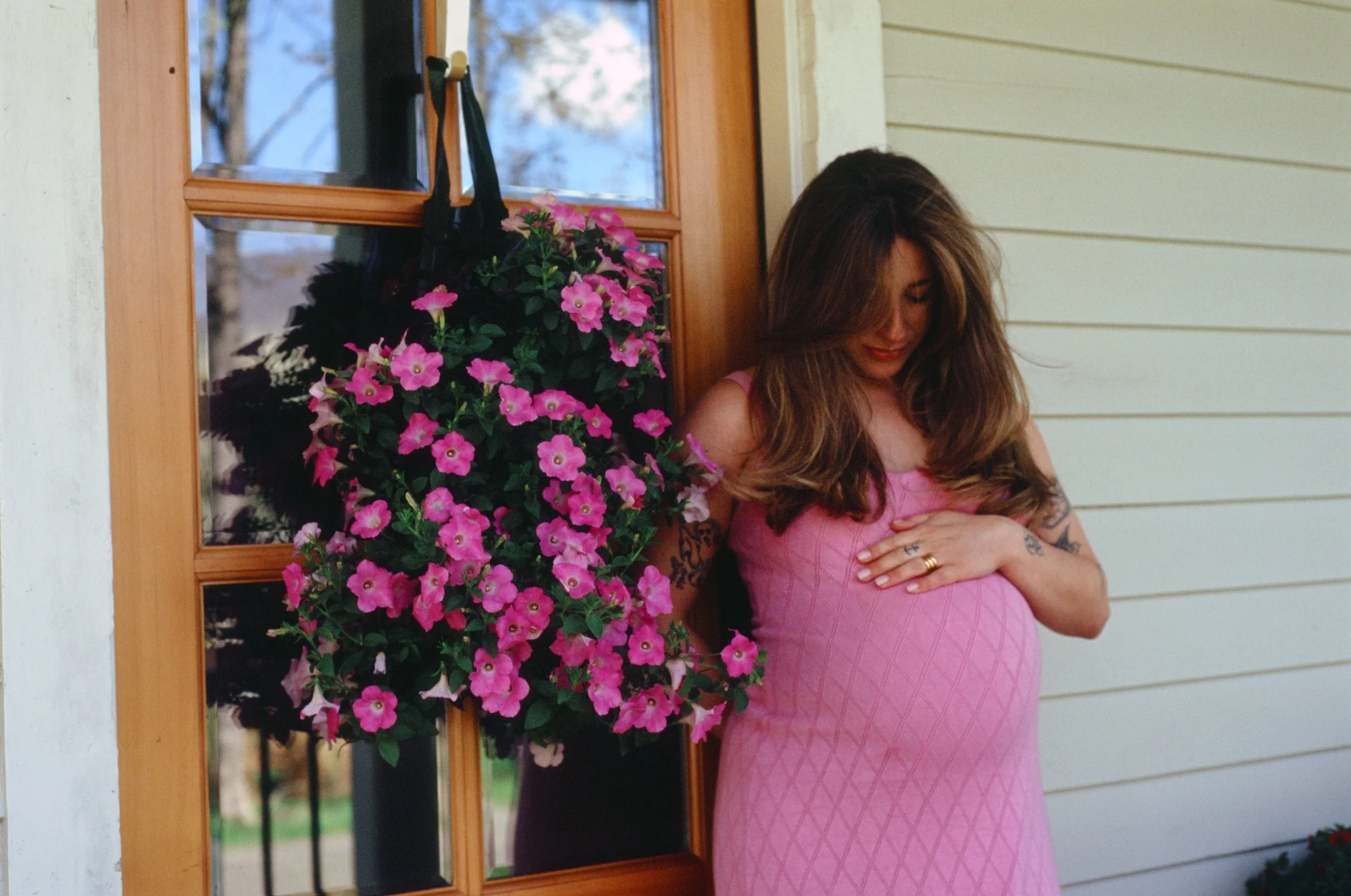 A woman with long brown hair, wearing a pink dress, is standing outside near a door with glass panels. She has her hand on her stomach and is smiling. Next to her is a large pink flowering plant hanging on a black hook on the door.