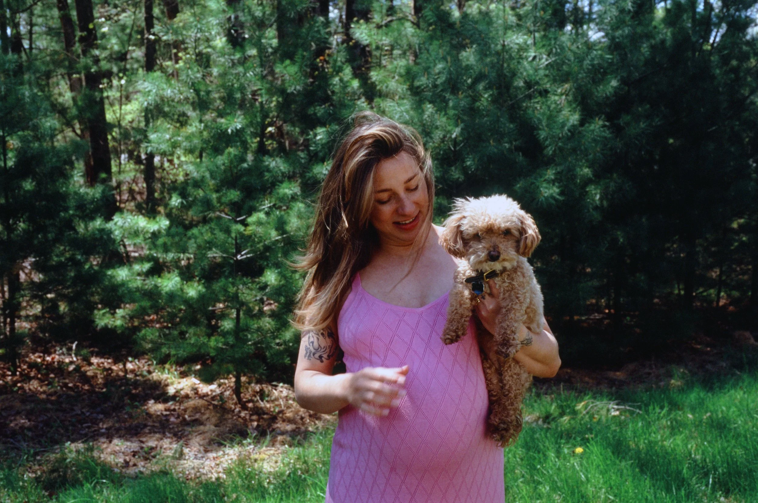 A woman with long hair holding a small tan poodle dog in a park with green trees in the background.