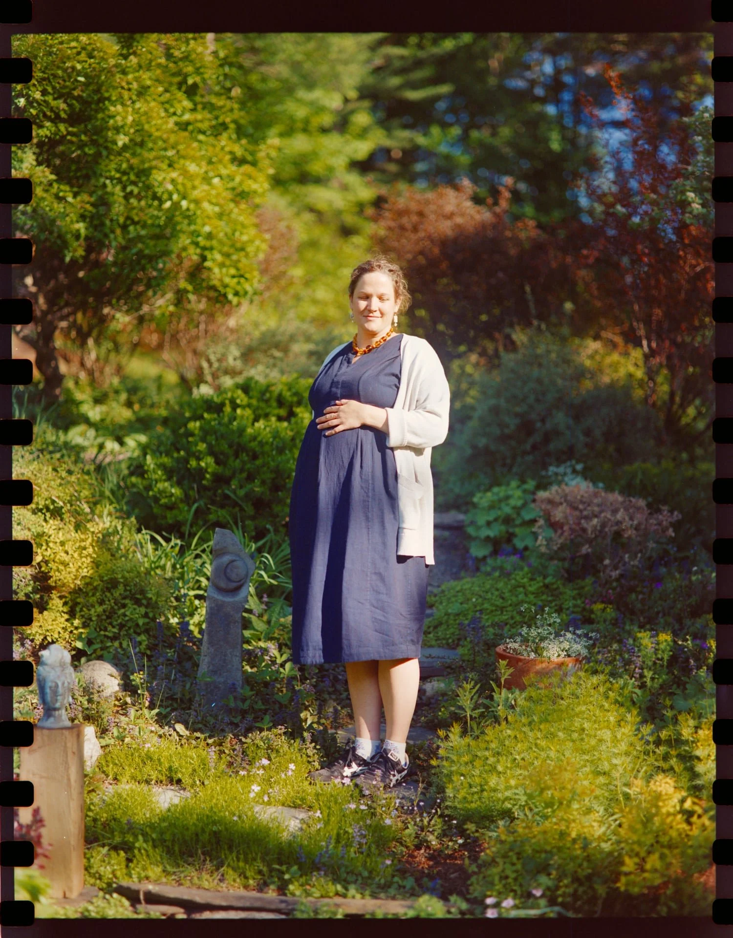 A woman standing in a lush garden with colorful trees and plants, wearing a dark dress, white cardigan, sneakers, and a necklace, with her hand on her stomach and eyes closed. Maternity photograph. 