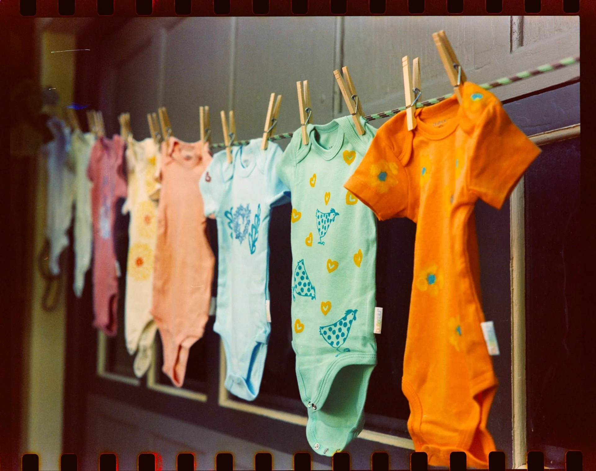 Colorful baby onesies hanging on a clothesline, attached with clothespins in front of a gray wall.