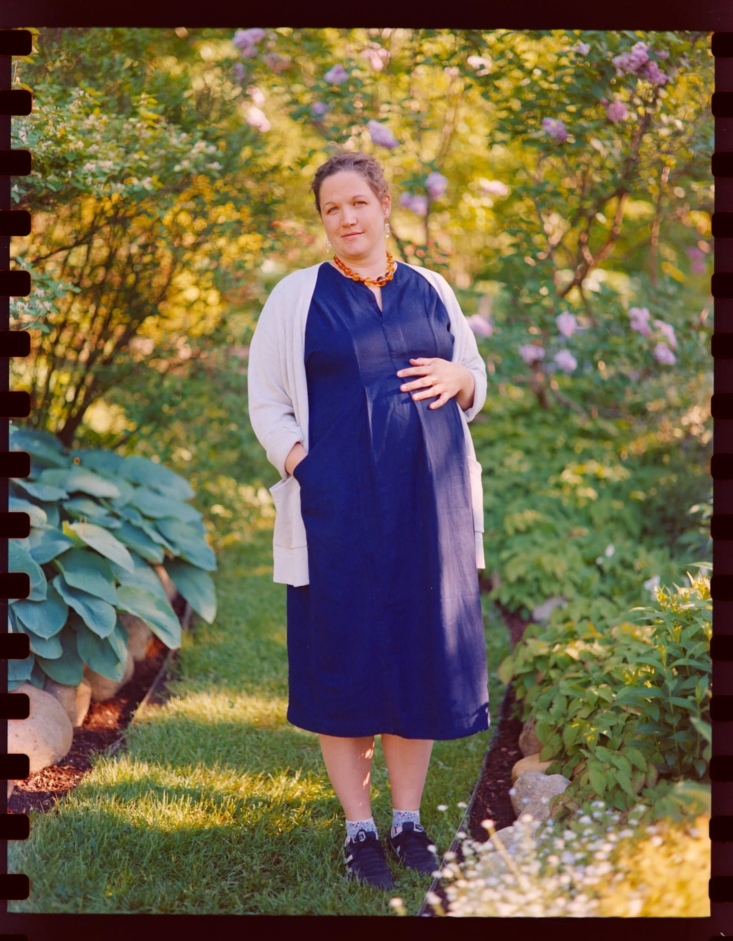 A woman standing in a garden with purple and pink flowers in the background, wearing a navy dress, white cardigan, and black sneakers.
