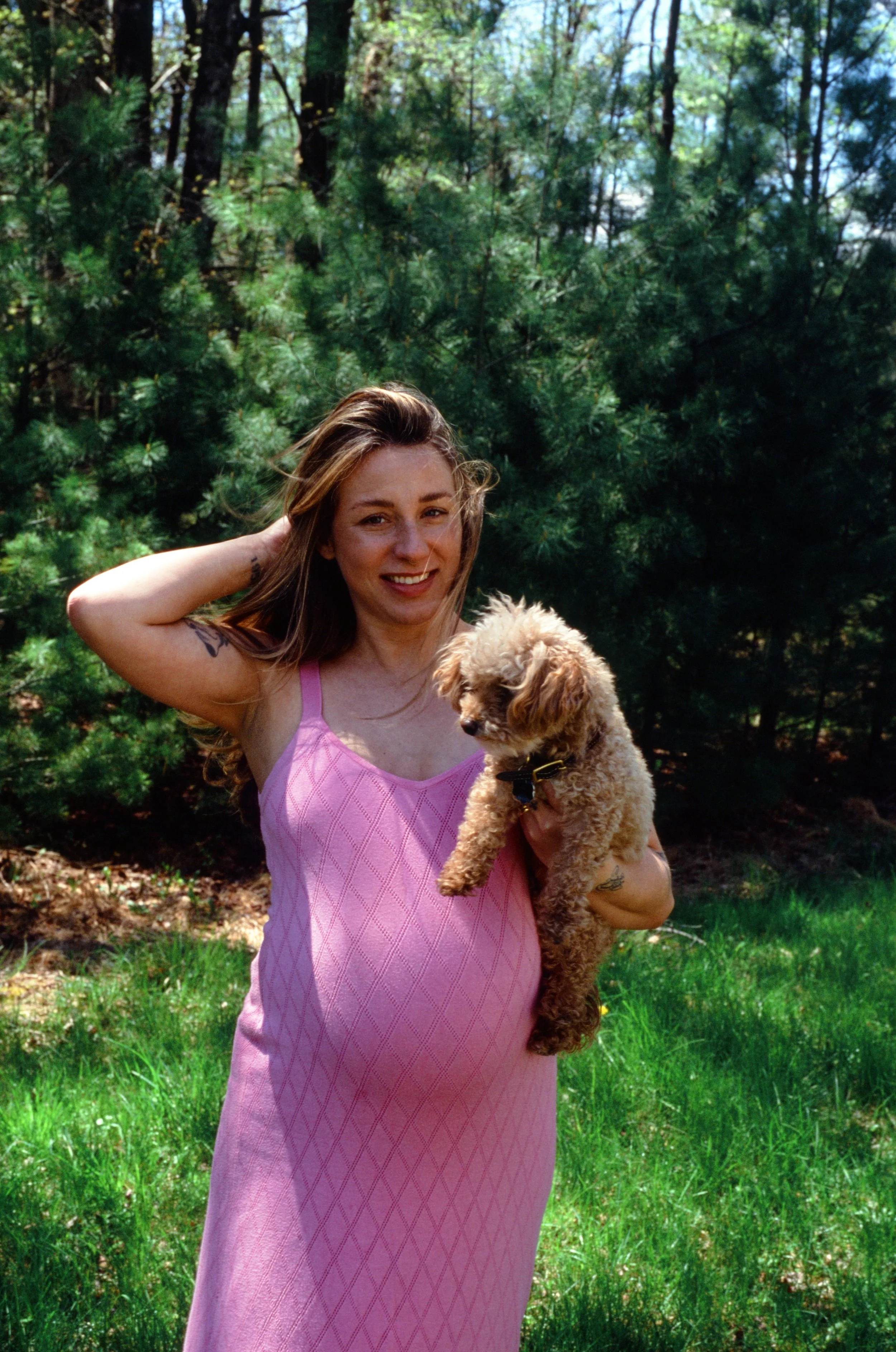 A woman with long hair wearing a pink dress holding a small, curly-haired dog in a green outdoor park or forest setting.