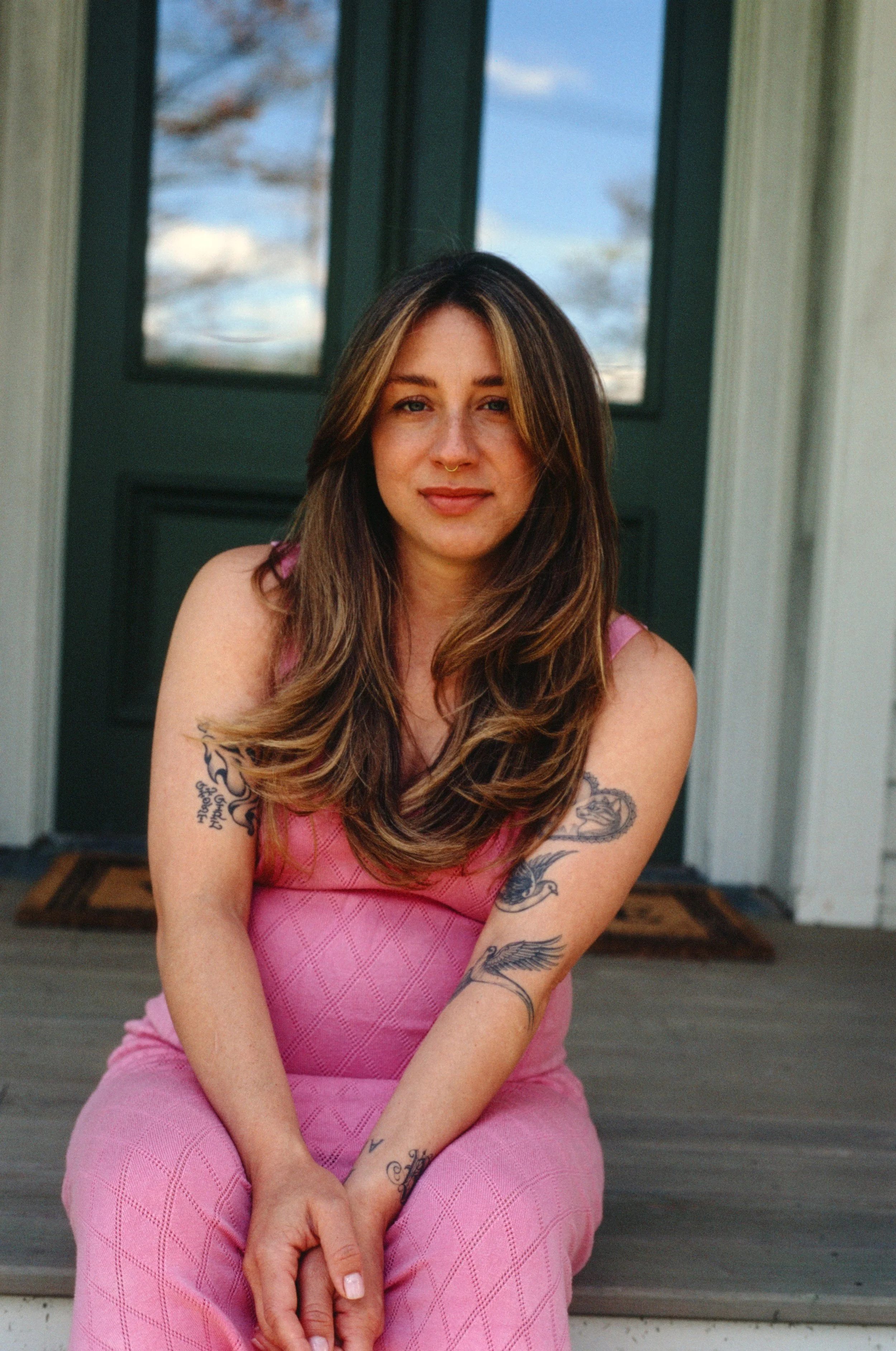 A woman with long brown hair, tattoos, and a septum piercing, wearing a pink dress, sitting on front steps in front of a green door with glass panes, outdoors during daytime.