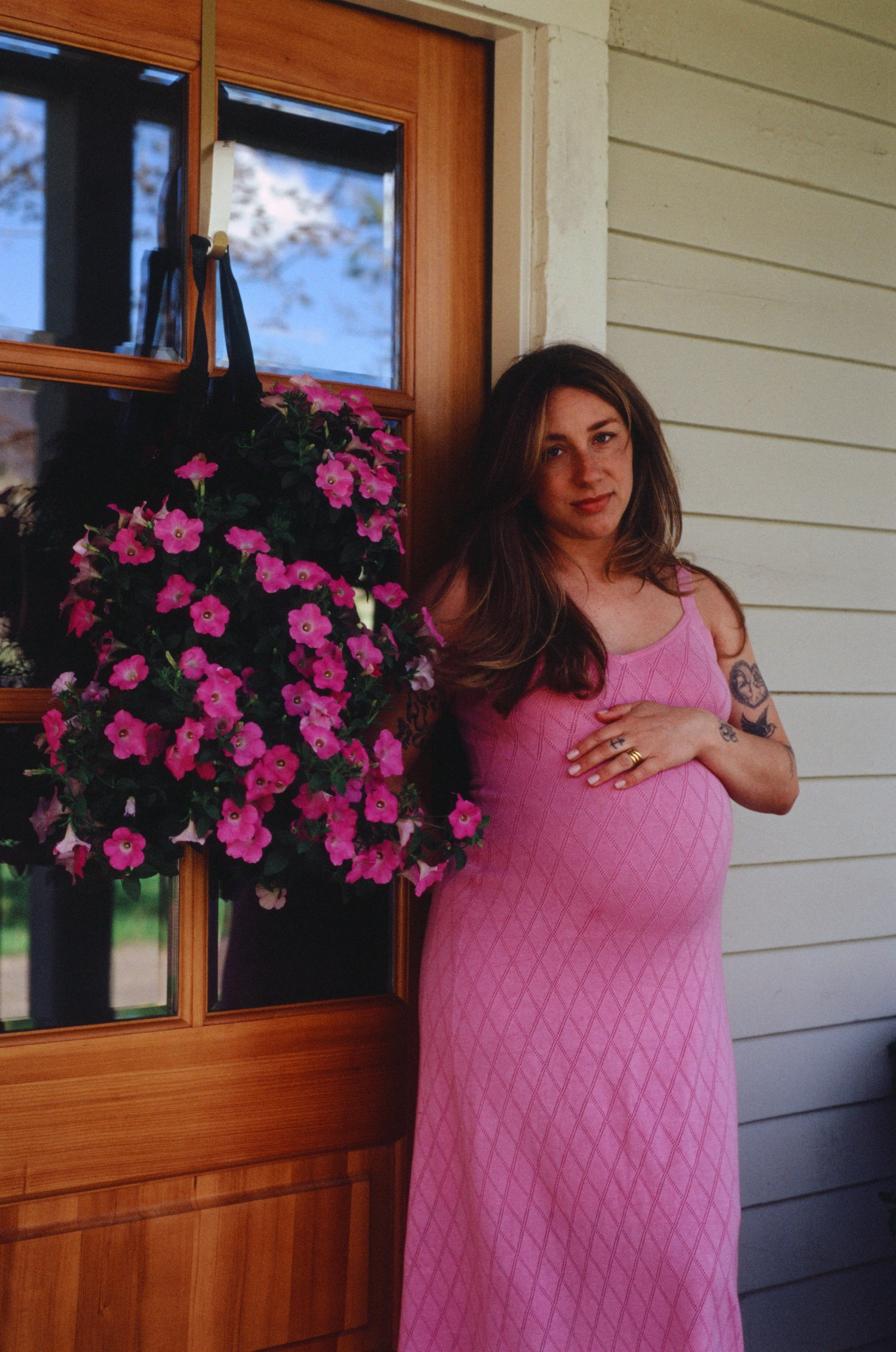 A pregnant woman with tattoos on her arms, wearing a pink sleeveless dress, standing in front of a wooden window with two panes of glass, next to a hanging basket of pink flowers.