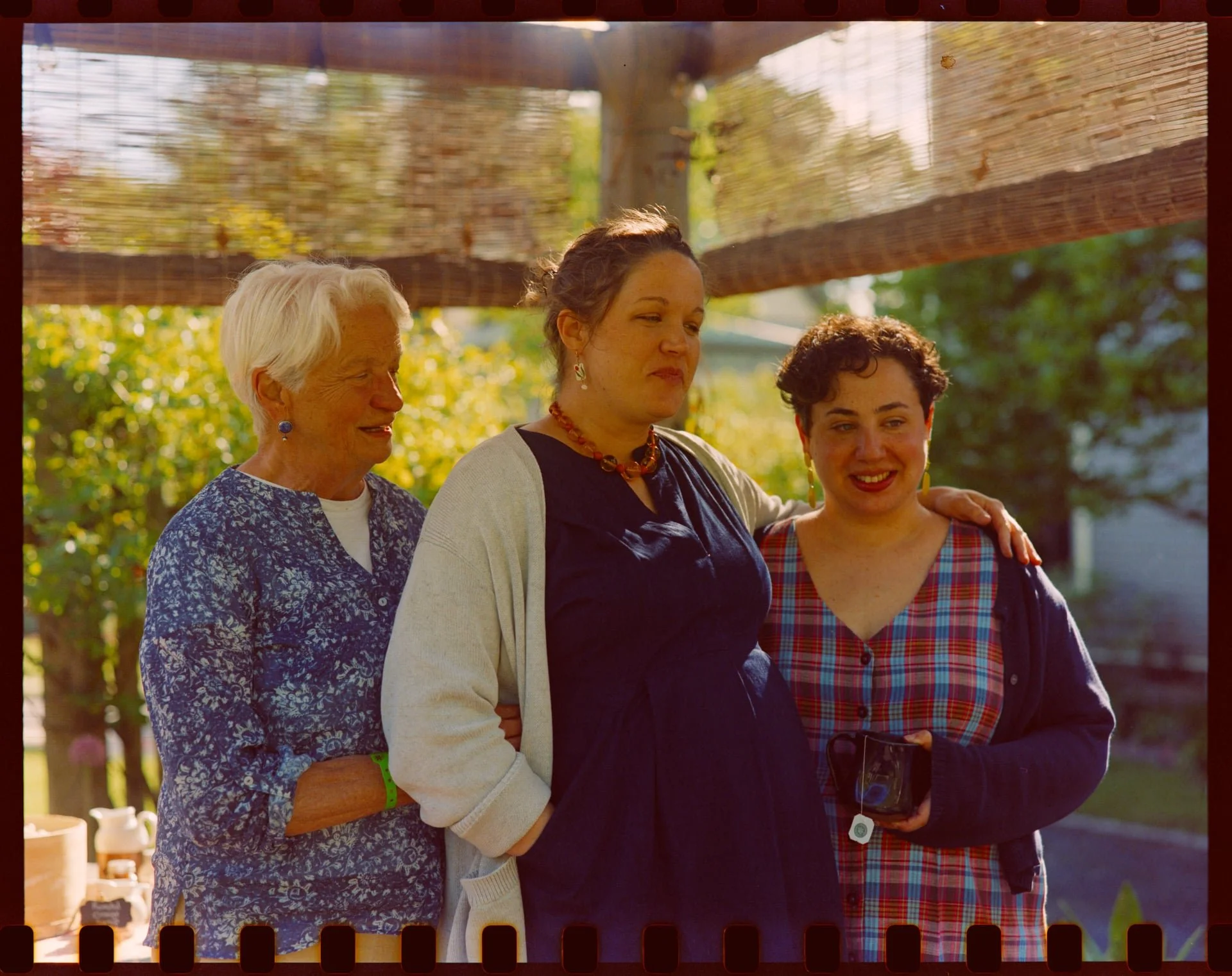 Four women standing outdoors under a wooden shelter, smiling and interacting with each other on a sunny day. There are trees and a house in the background.