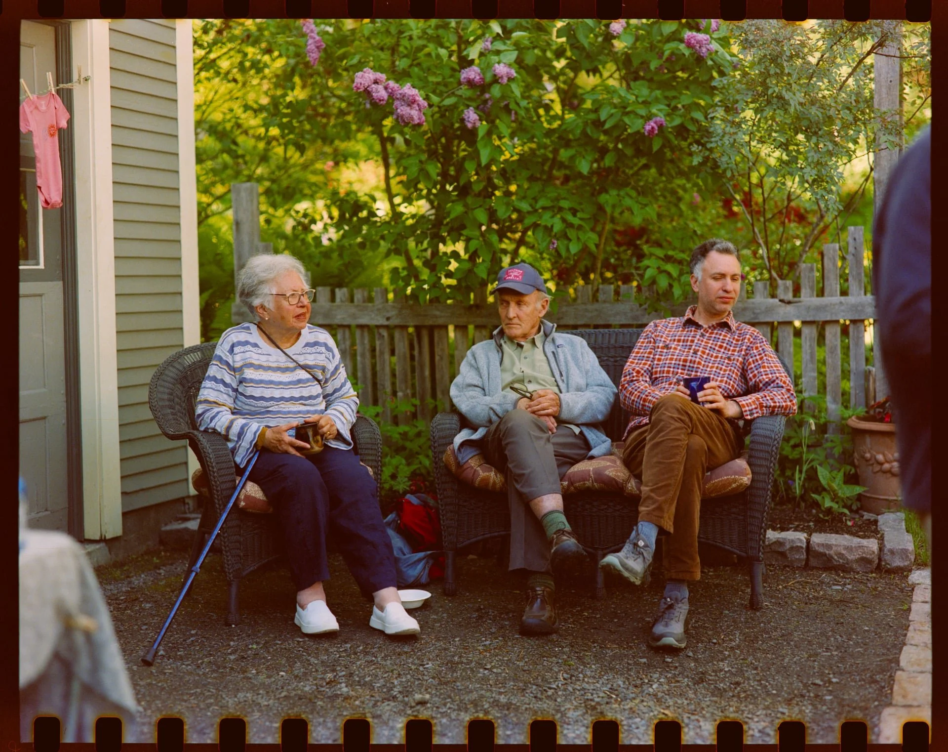 Three elderly people sitting in outdoor chairs in a backyard, with lush green trees and purple flowers in the background. One woman, wearing glasses and a striped shirt, is holding a mug. An elderly man, wearing a baseball cap and gray cardigan, is s