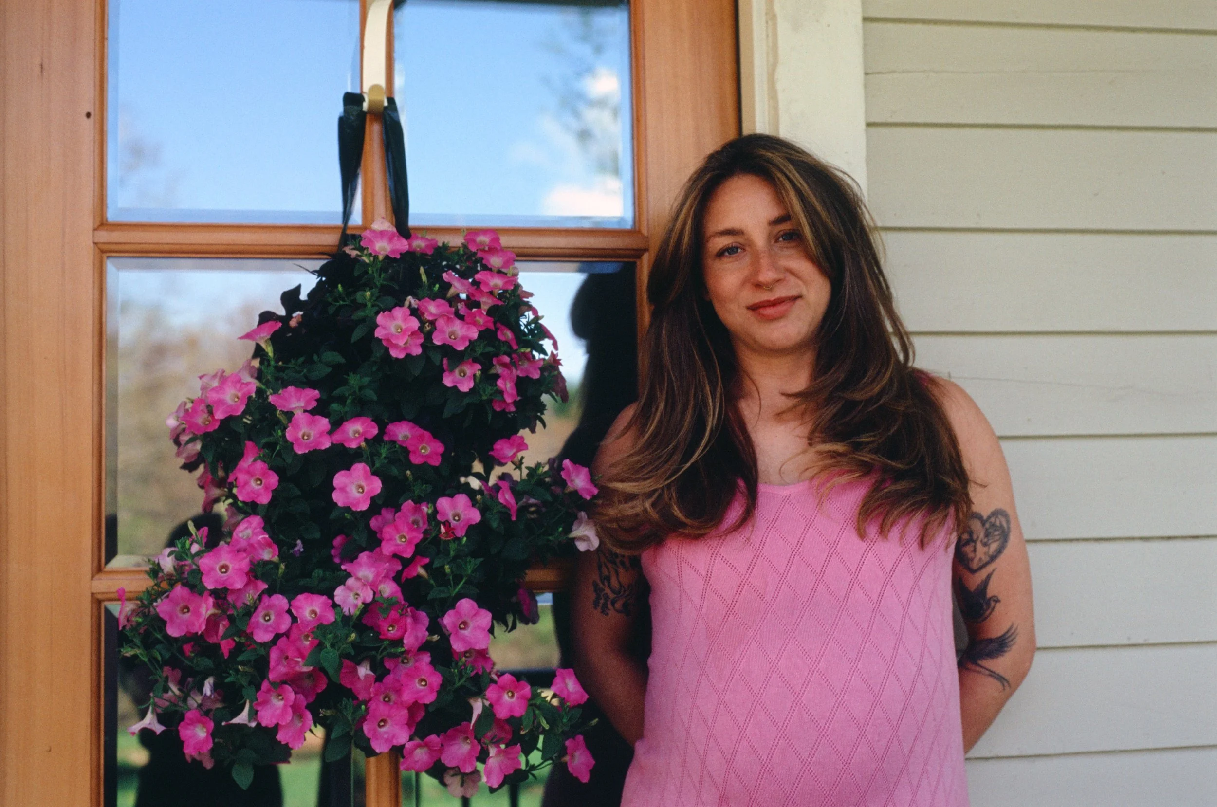 A woman with long brown hair and tattoos on her arms, wearing a pink dress, standing next to a large pink flowering plant in front of a wooden window and beige house siding.