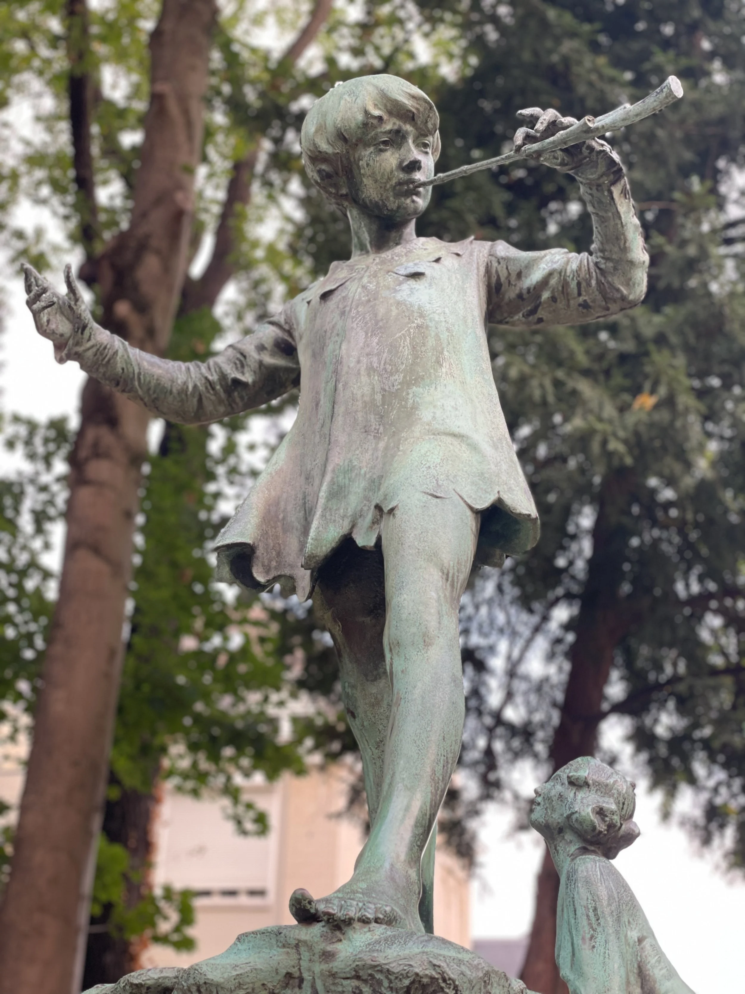 Bronze statue of a young child playing a flute, standing on a rock with a dog at their feet, surrounded by trees.
