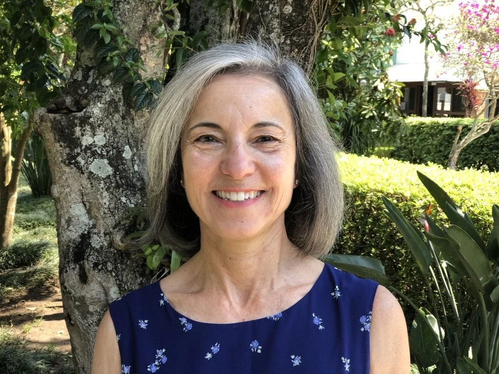 A smiling woman with gray hair, wearing a navy blue dress with small white floral patterns, outdoors in a garden with trees and bushes in the background.