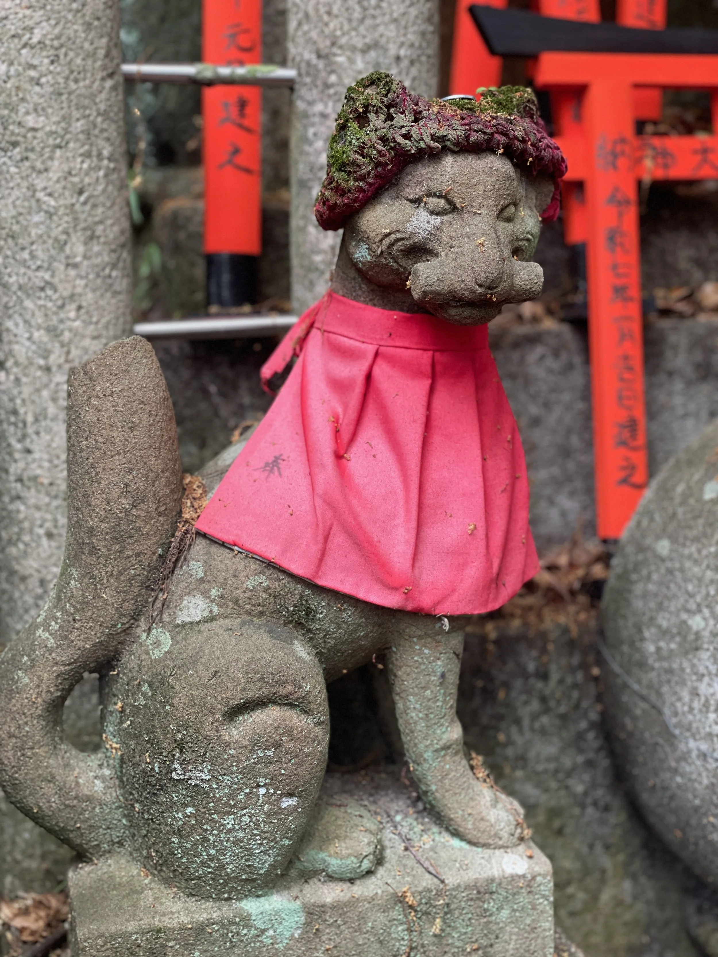 A small stone statue of a dog with a human-like face, adorned with a pink cloth around its neck, standing on a stone base. In the background, there are red and black Japanese or Chinese prayer plaques attached to a tree.