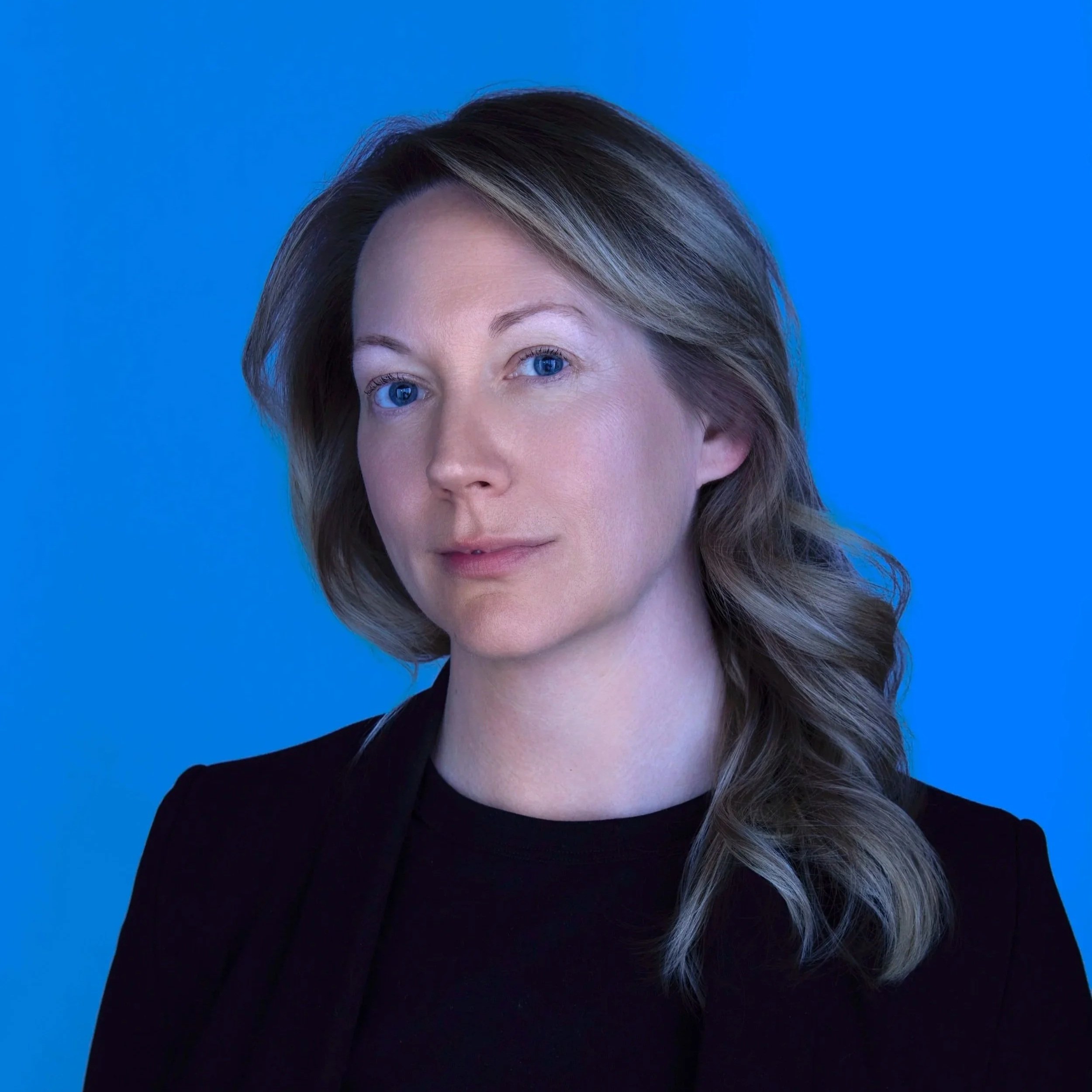 A portrait of a woman with shoulder-length wavy brown hair and blue eyes, wearing a black top and blazer, against a solid blue background.