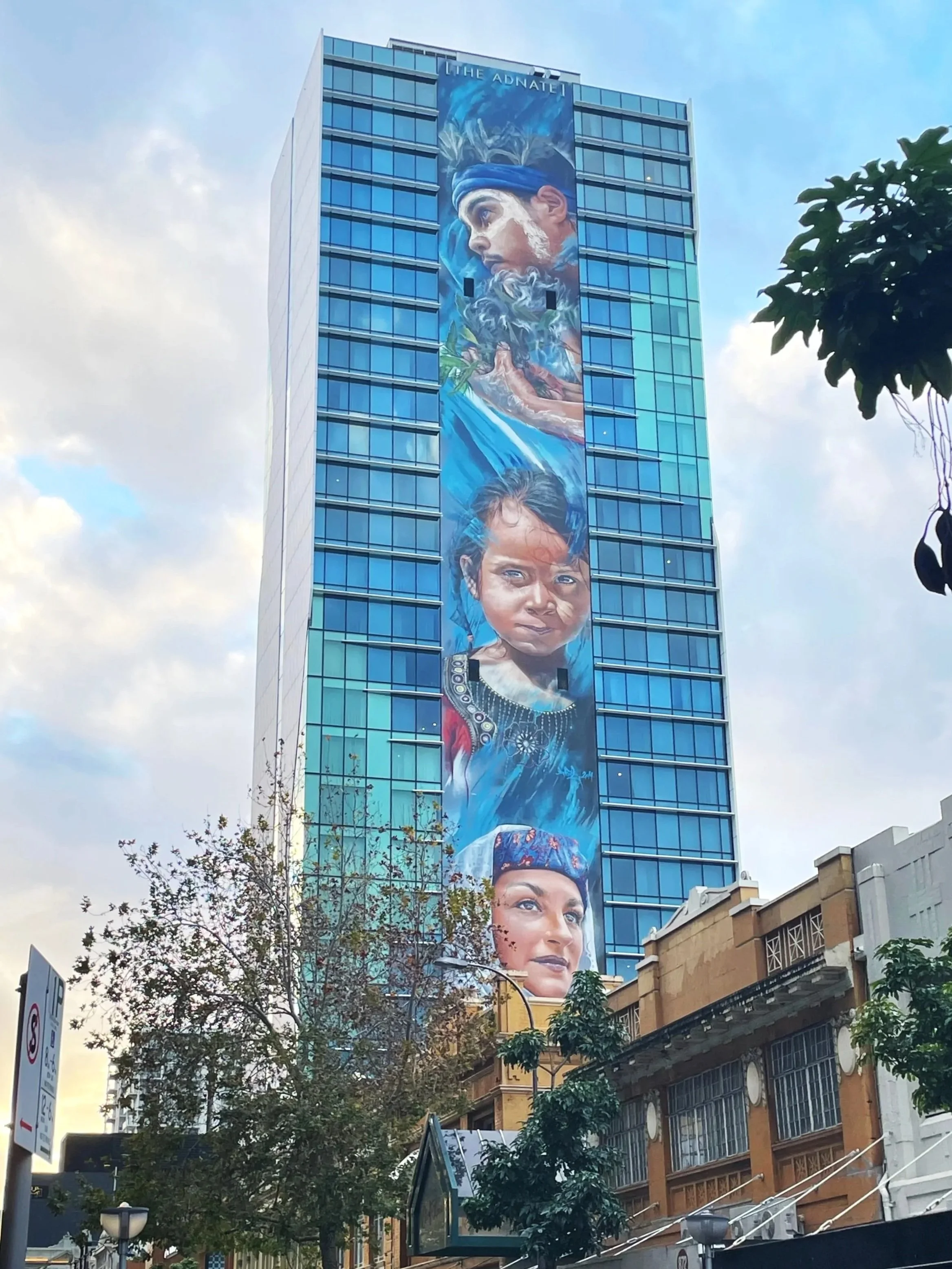 Large mural of diverse people's faces painted on a tall blue glass building, featuring a tribal woman, a young girl, a woman with a headscarf, and a bearded man with a headband, among clouds and water motifs.