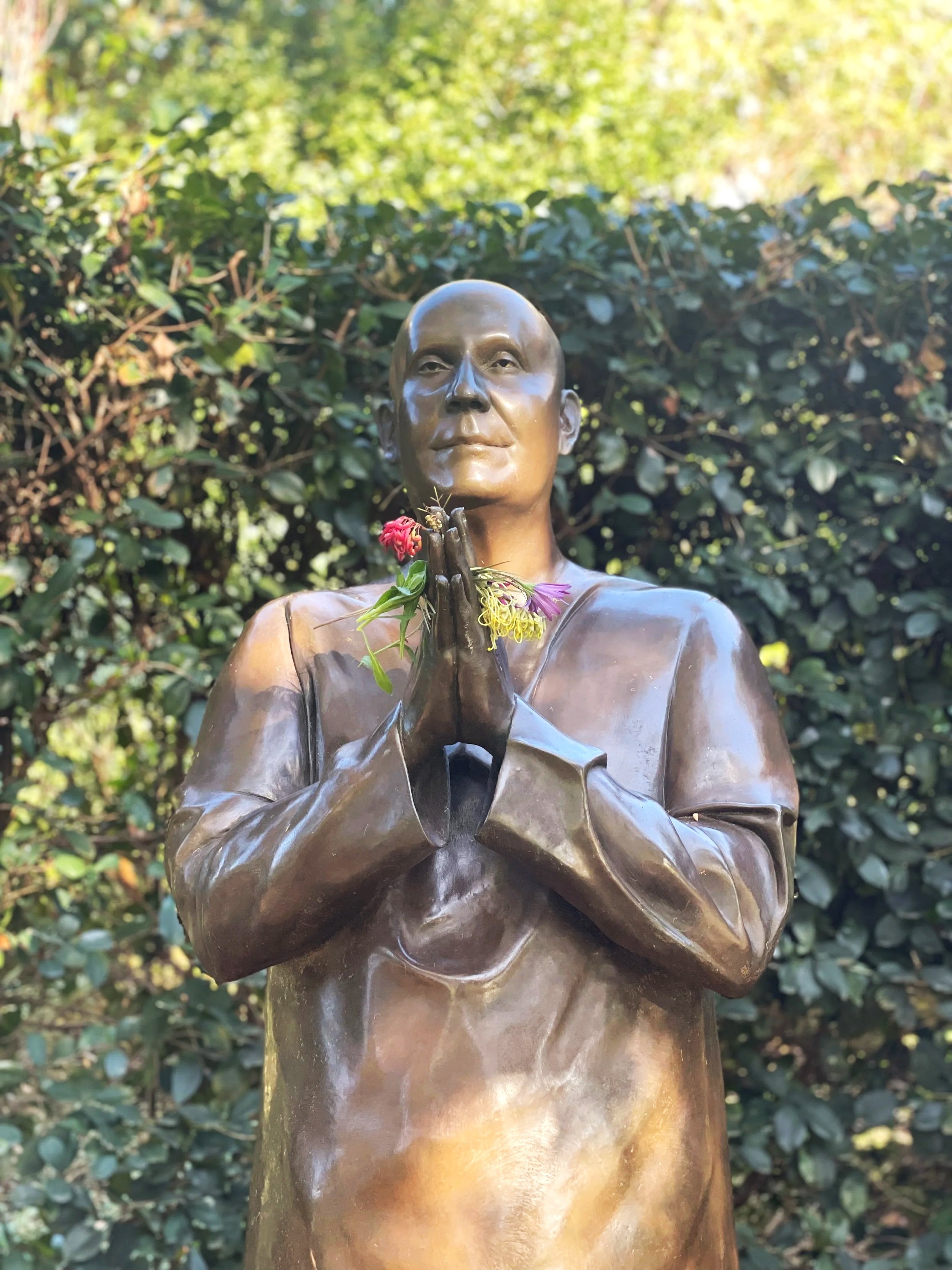 Bronze statue of a person with hands clasped in prayer, holding colorful flowers, outdoors with greenery in the background.