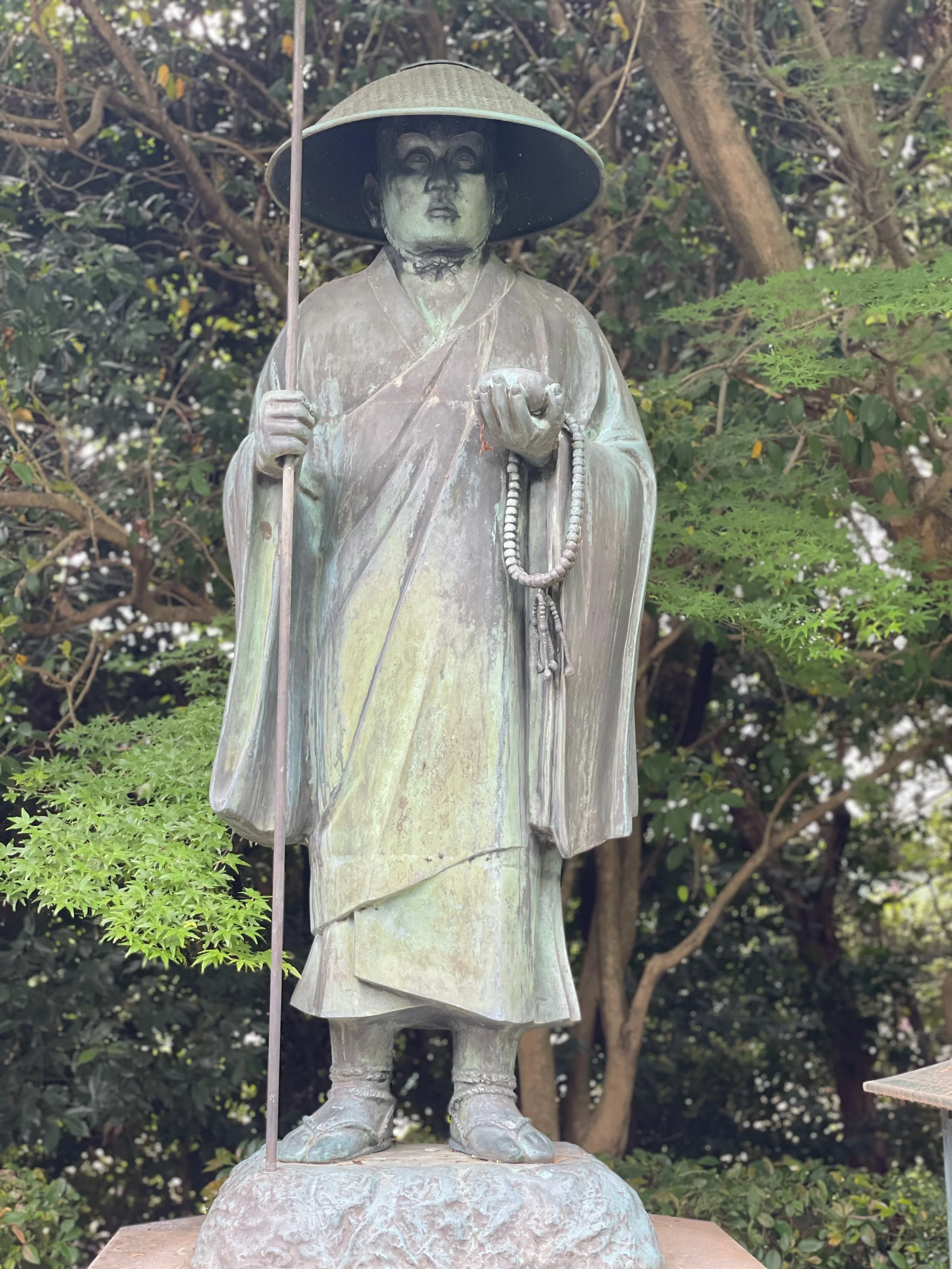 Bronze statue of a standing monk or religious figure wearing a large conical hat, holding a staff in one hand and prayer beads in the other, set outdoors with trees and greenery in the background.