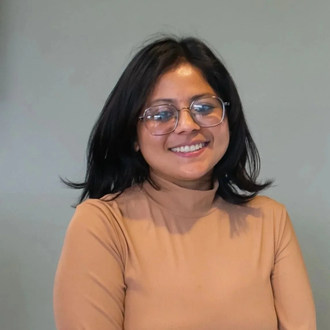 A young woman with shoulder-length black hair, glasses, and a beige turtleneck sweater, smiling against a plain gray wall.