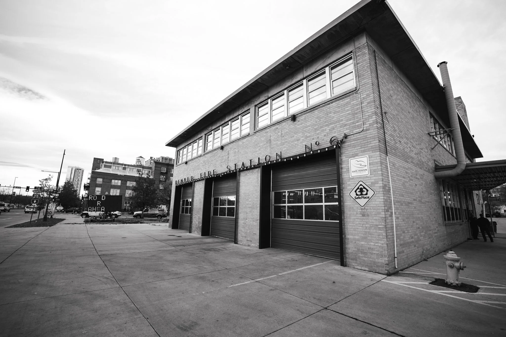 Black and white photo of the exterior of Denver Fire Station No. 6 with three garage doors, signs, and city buildings in the background. Concert Design Studio STATION SIX.