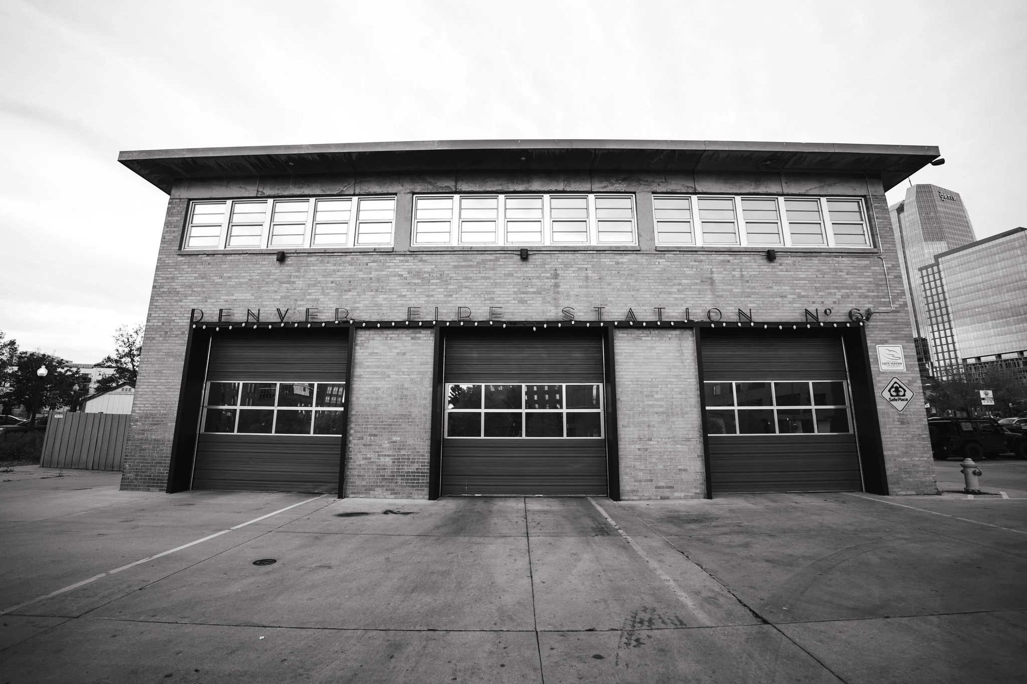 Black and white photo of the exterior of Denver Fire Station No. 6 with three garage doors, signs, and city buildings in the background. Concert Design Studio STATION SIX.