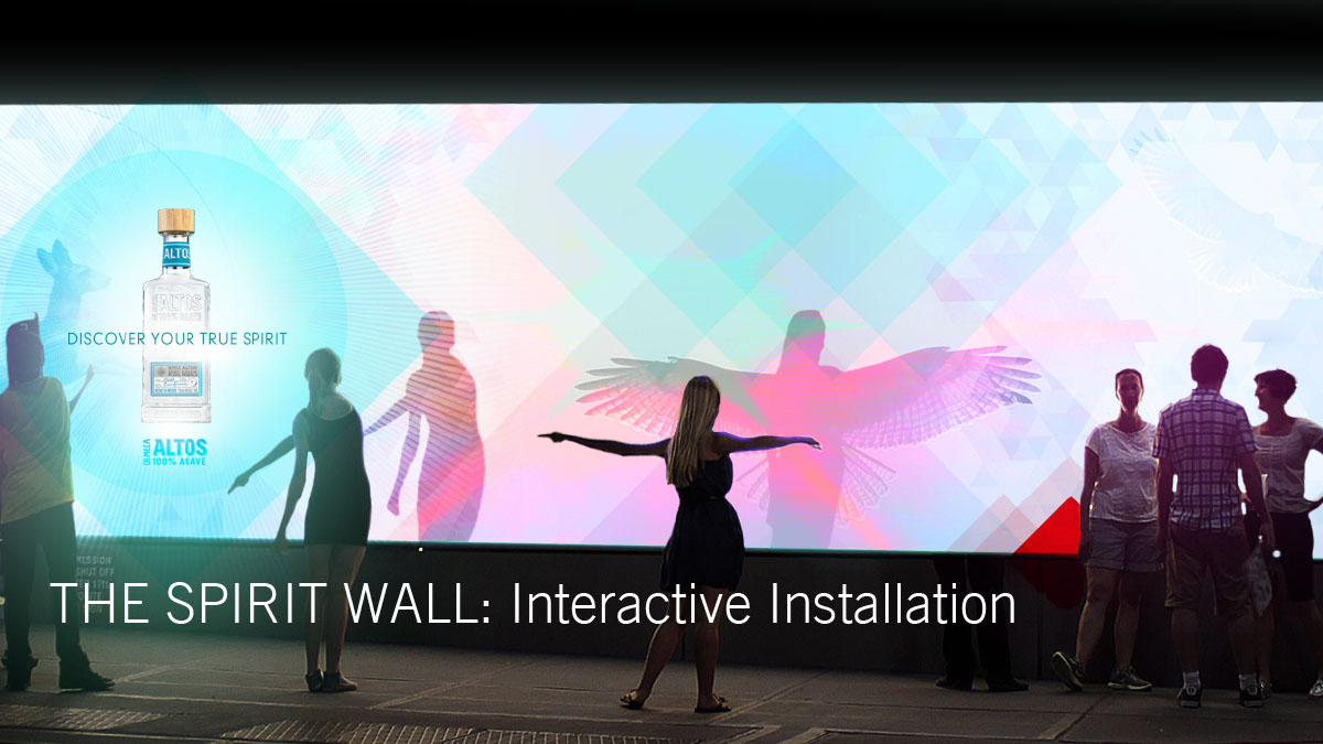 A large digital display called 'The Spirit Wall: Interactive Installation' showing graphics of a bottle, a butterfly, a person with outstretched arms, a person with wings, and a group of people standing and interacting.