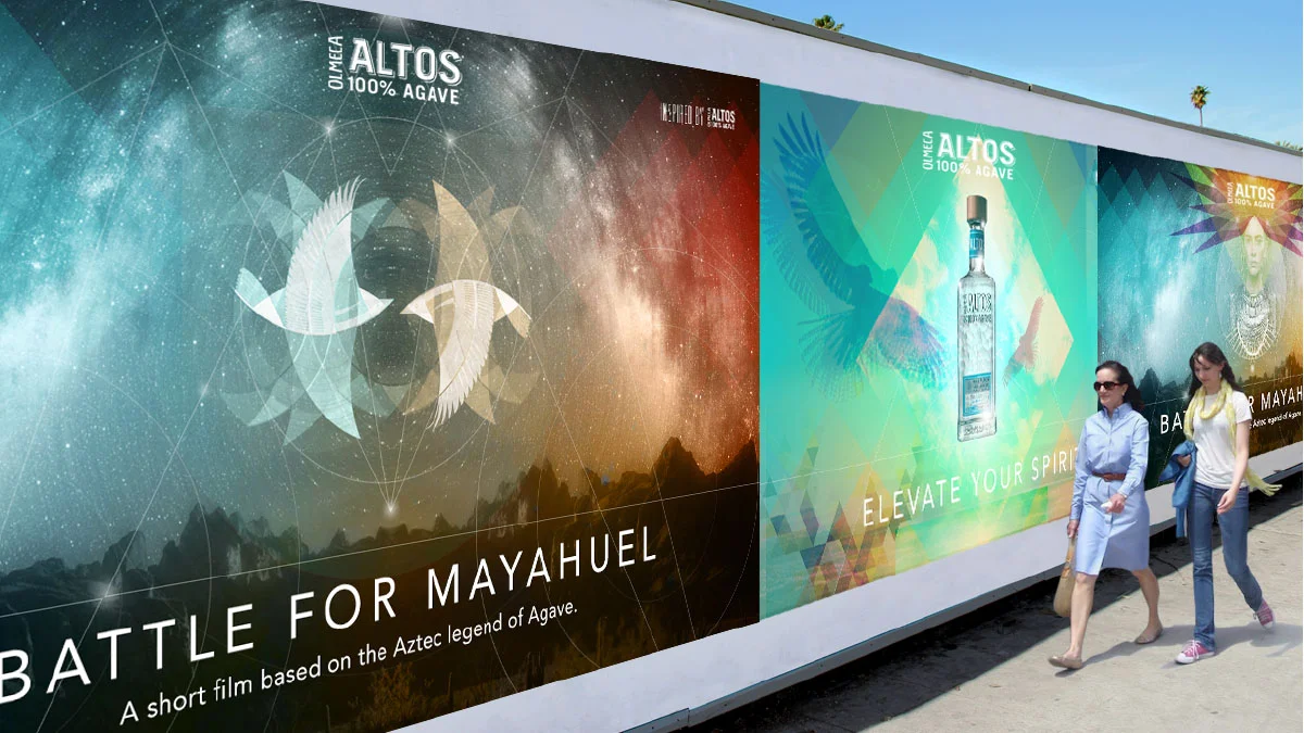 Large outdoor advertisement with colorful graphics on a wall. The left side features artwork of two stylized birds and stars, with the text "BATTLE FOR MAYAHUEL" and a subtitle about an Aztec legend of Agave. The right side displays an image of a bot
