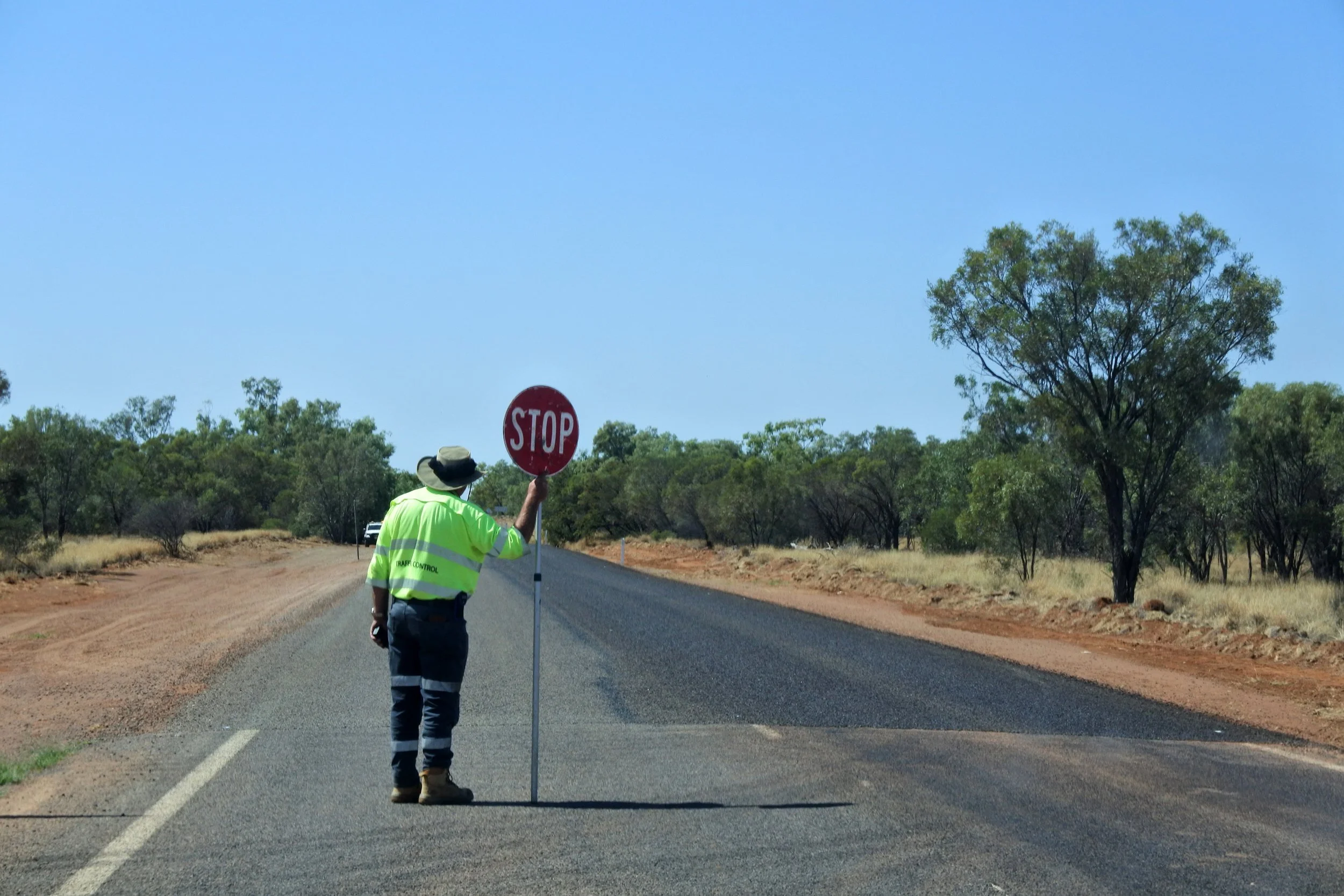 Road worker holding a STOP sign on a rural road during daytime, wearing a high-vis shirt and wide-brim hat, with trees and dry roadside vegetation in the background.