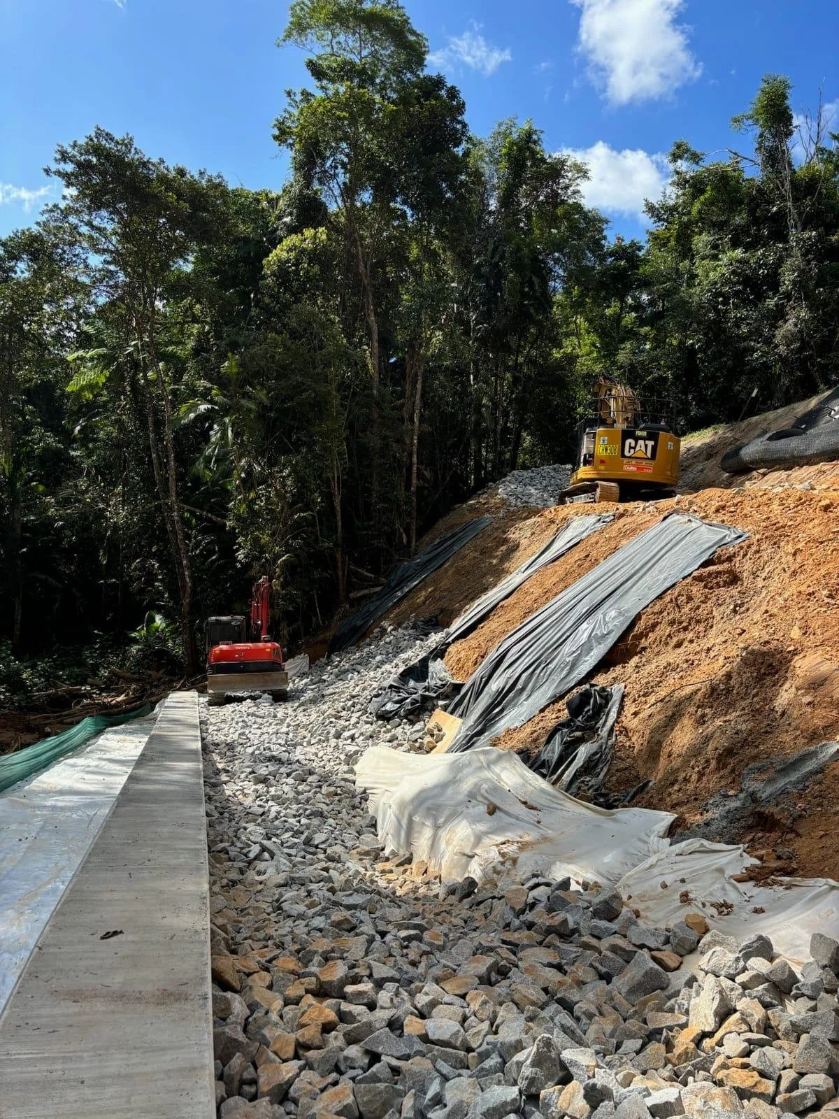 Civil works underway on a steep embankment, with excavators, rock scour protection and erosion control materials installed along a rainforest hillside.