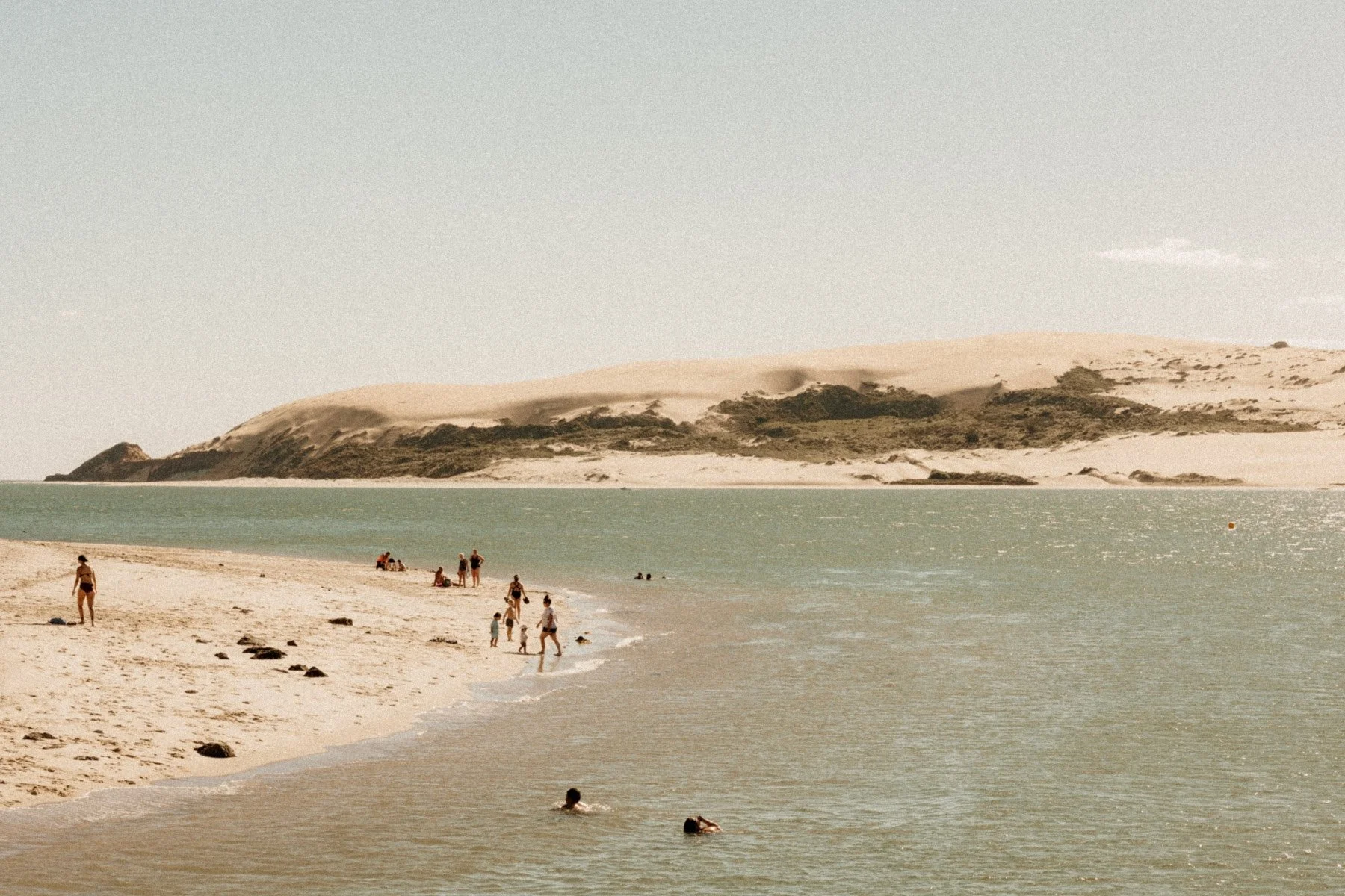 Beach with people swimming and relaxing, sandy shore, dunes in the background under a clear sky.