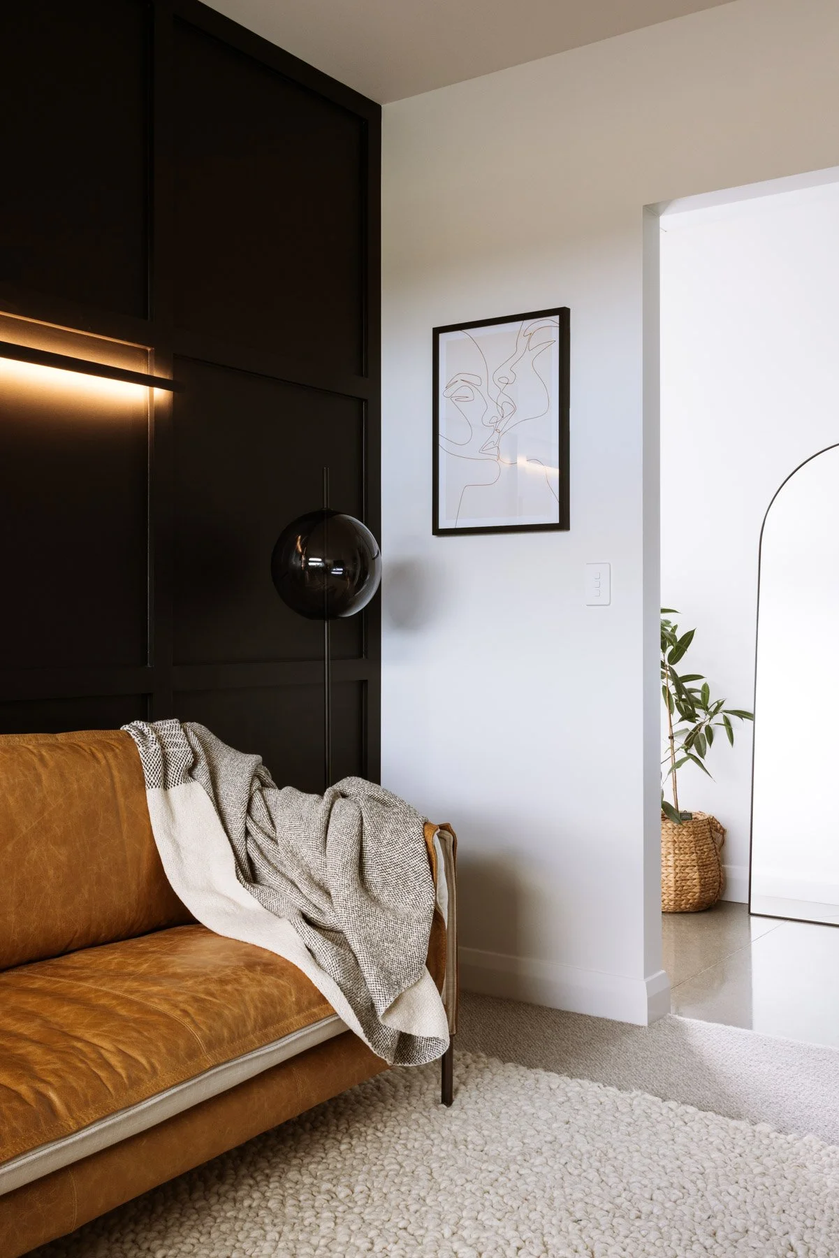 Living room with tan leather couch, black wall panel, black spherical floor lamp, abstract line art picture, potted plant, and cream-colored textured rug.