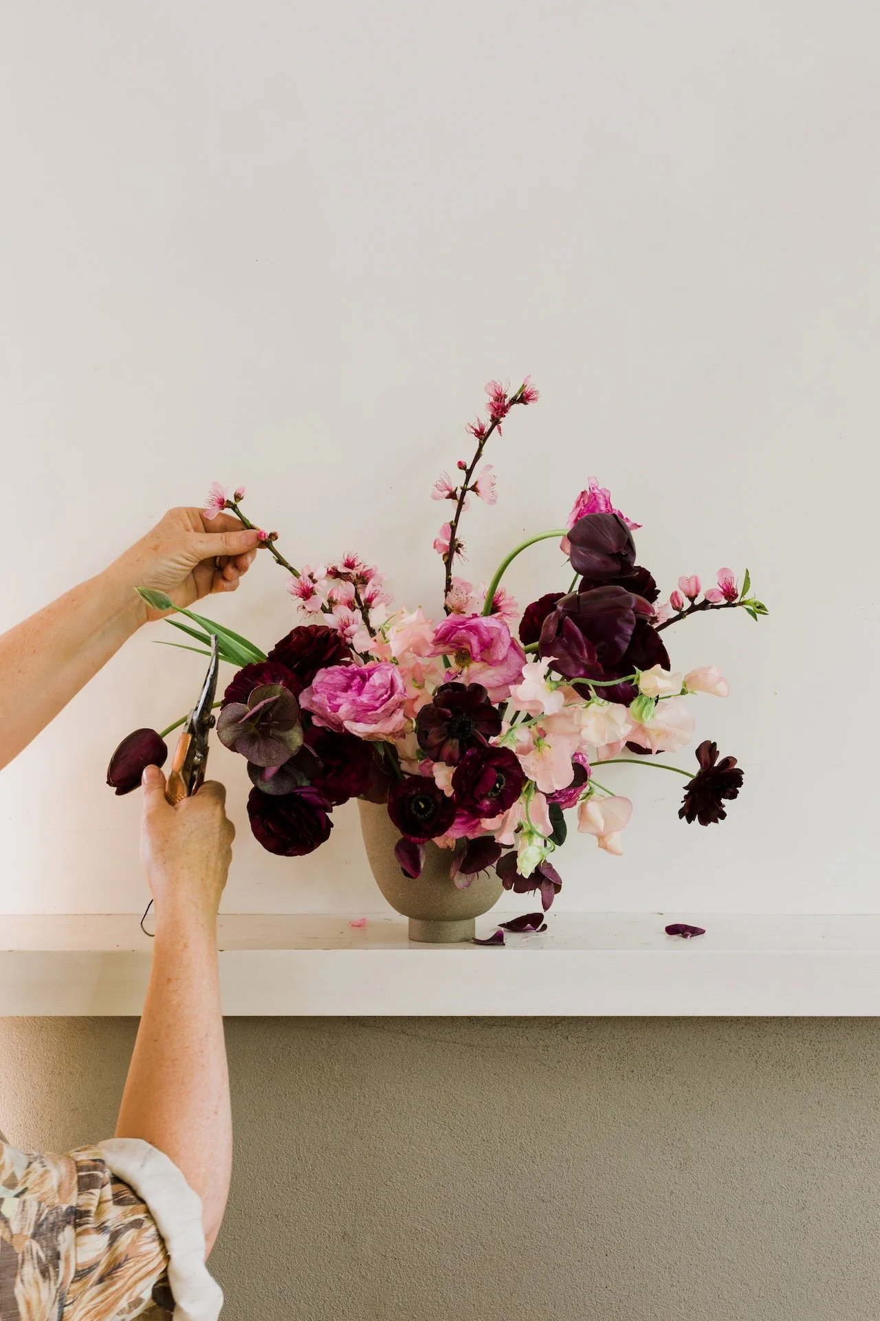 A person arranging a bouquet of pink and dark purple flowers in a gray vase on a white shelf. Lifestyle branding photography.