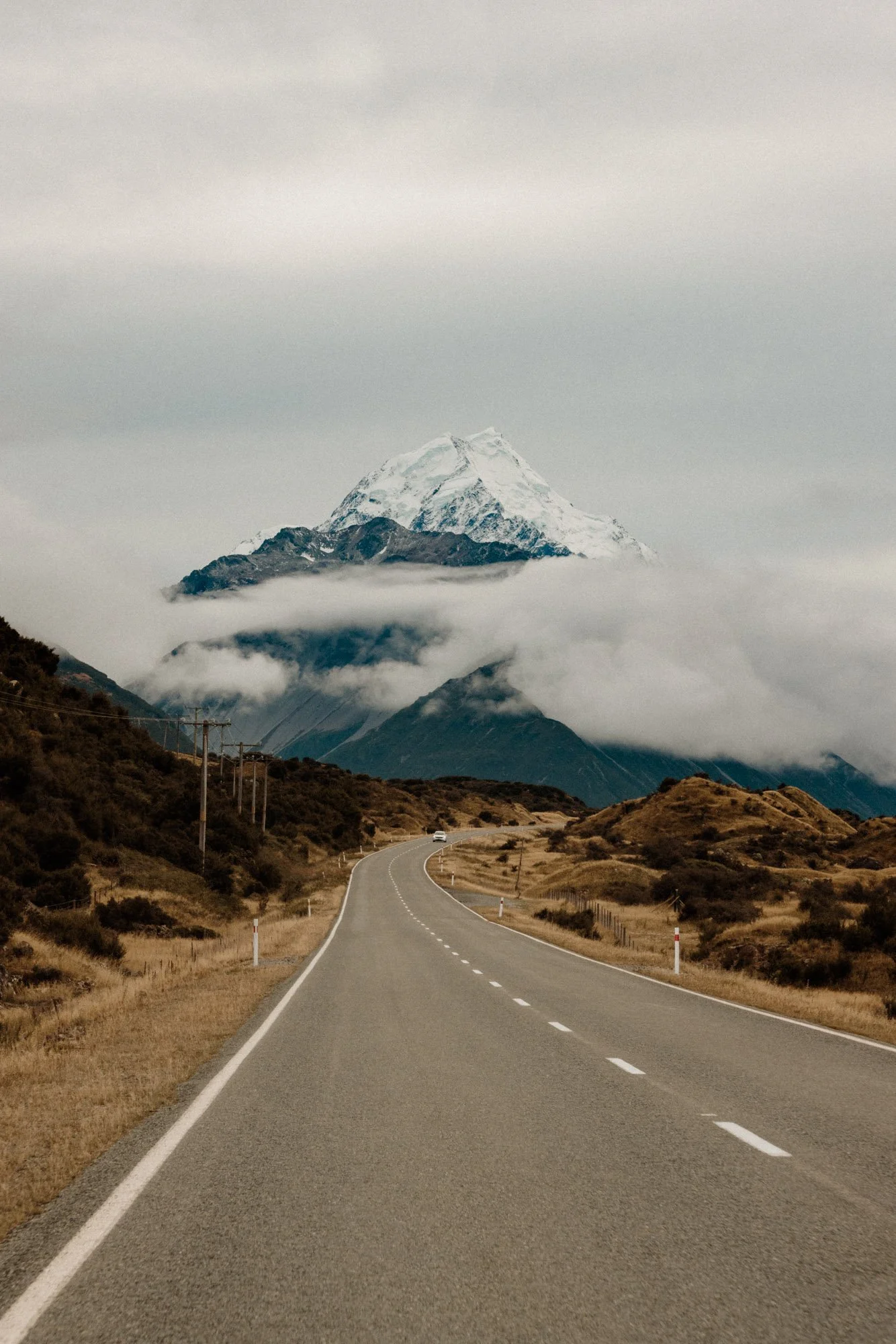A winding road leading towards snow-capped mountain with clouds around its base and on the sky.
