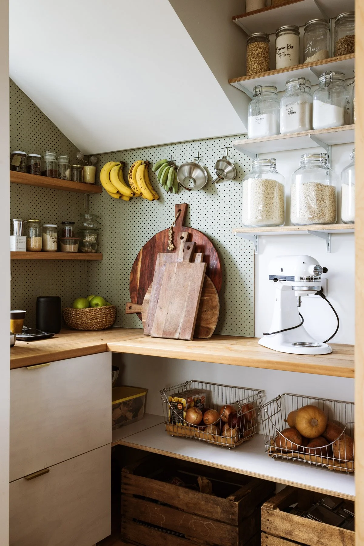 A tidy kitchen corner with wooden shelves holding jars of flour, sugar, and grains, a bunch of bananas and green apples hanging on the wall, cutting boards leaning against a pegboard, a white stand mixer, baskets of onions and potatoes, and a few box
