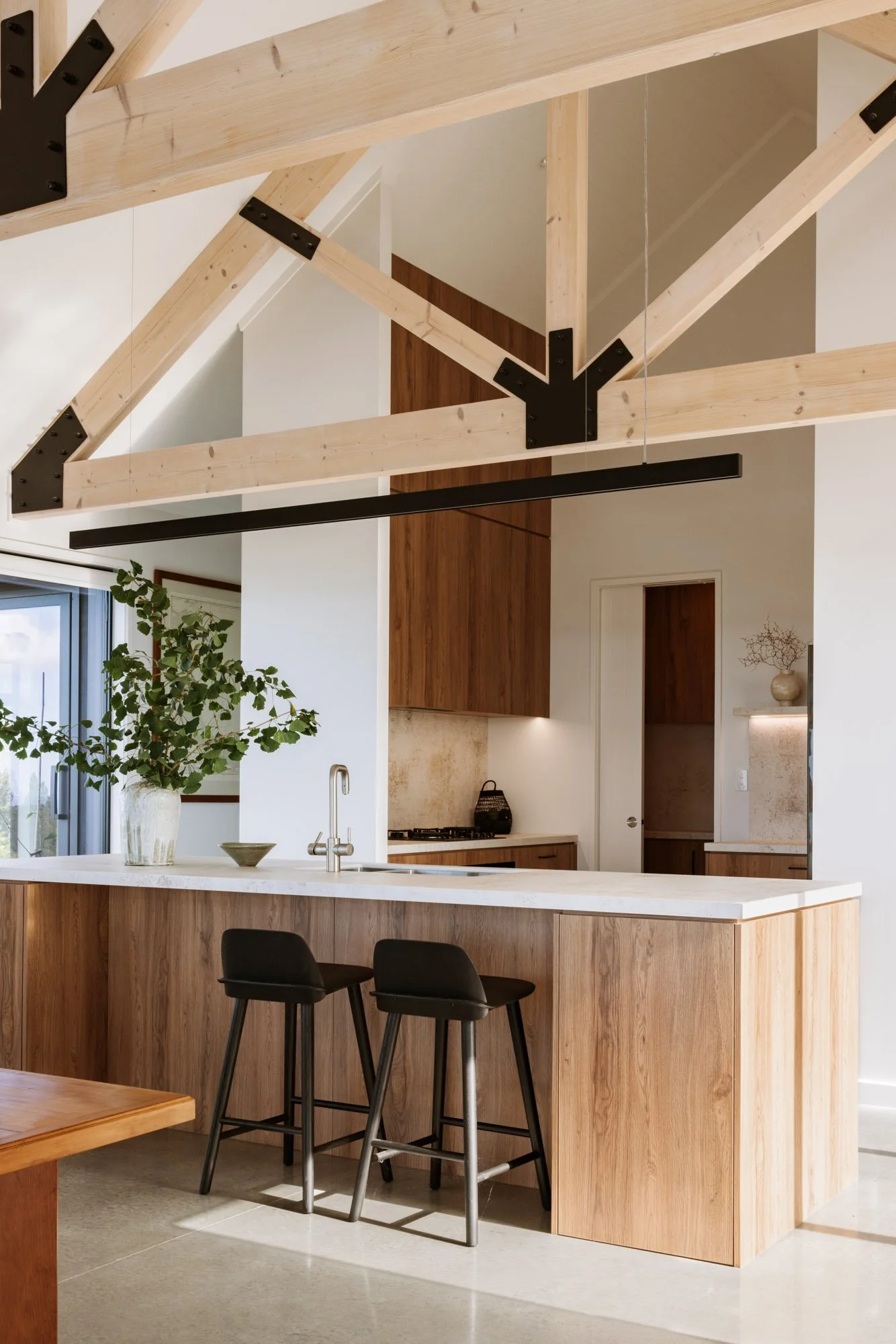 Modern kitchen with wooden cabinets and a white countertop, featuring a large window with greenery outside. A wooden mezzanine with black metal supports hangs above, and two black bar stools are at the kitchen counter.