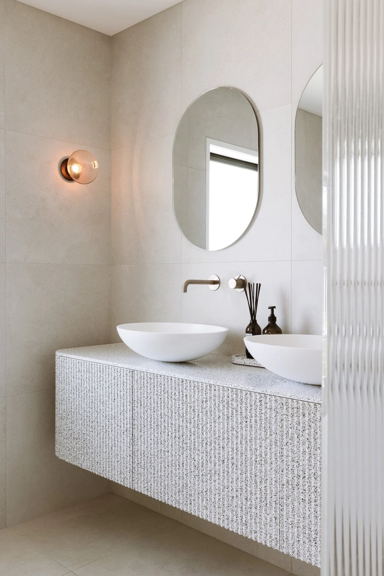 Modern bathroom with a floating textured vanity, two white vessel sinks, oval mirrors, a wall-mounted faucet, black and clear decor bottles, and a round wall light fixture.