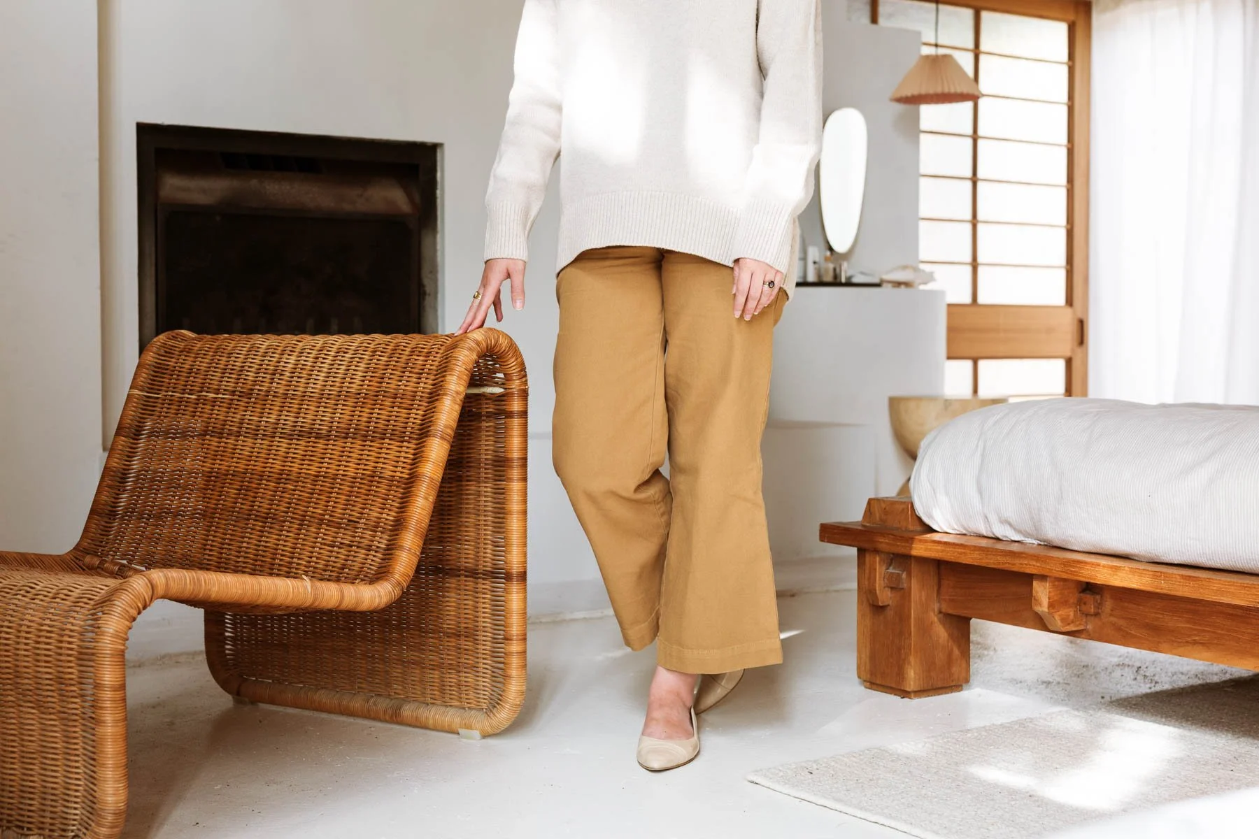 A person is standing in a cozy, minimalist bedroom, wearing a white sweater, tan wide-leg pants, and beige shoes, with one hand resting on a wicker chair. Lifestyle commercial branding photography in New Zealand.