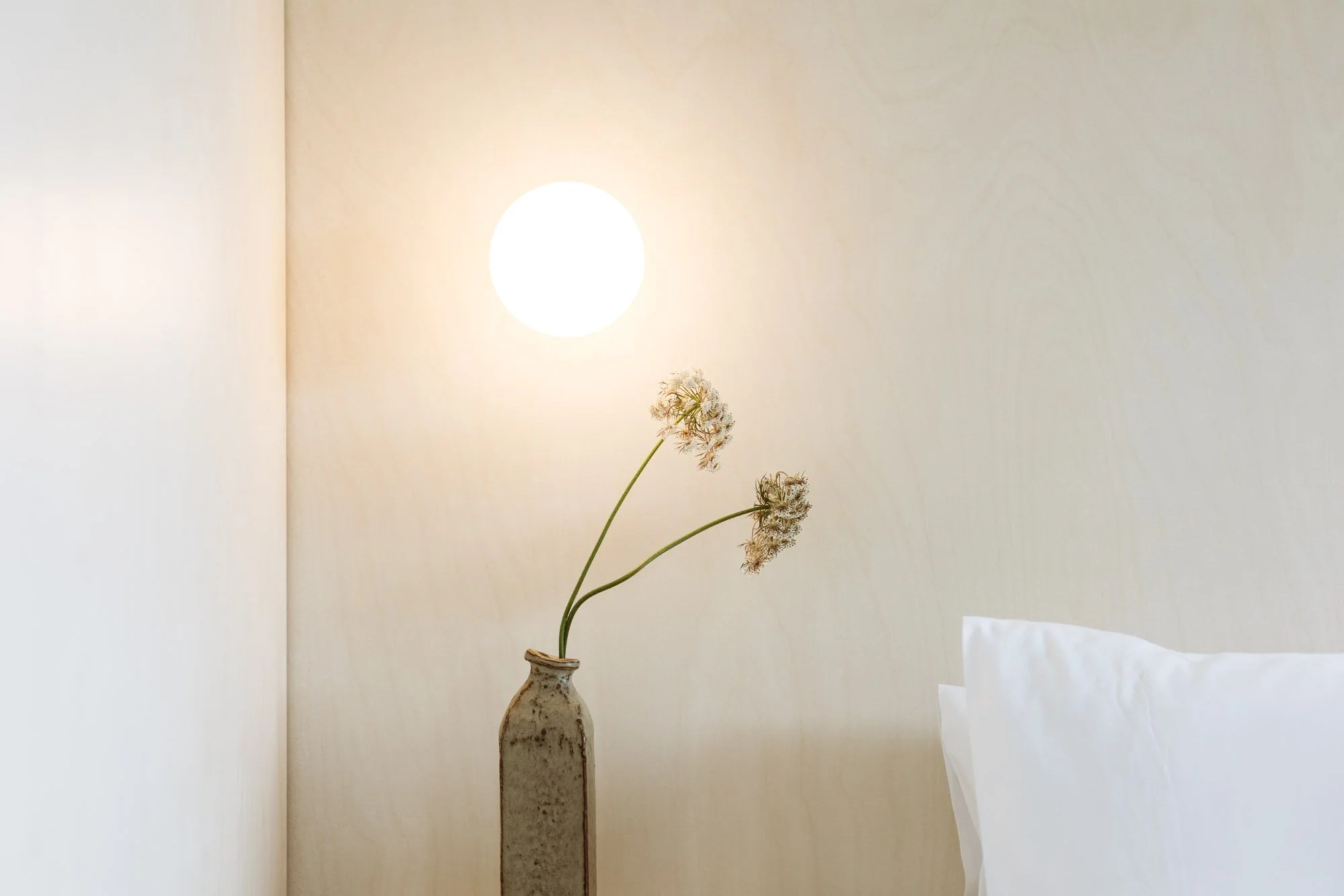 A minimalist bedroom corner with two dried flowers in a tall ceramic vase, a wall-mounted round light, and a white pillow on a bed against a beige wall.